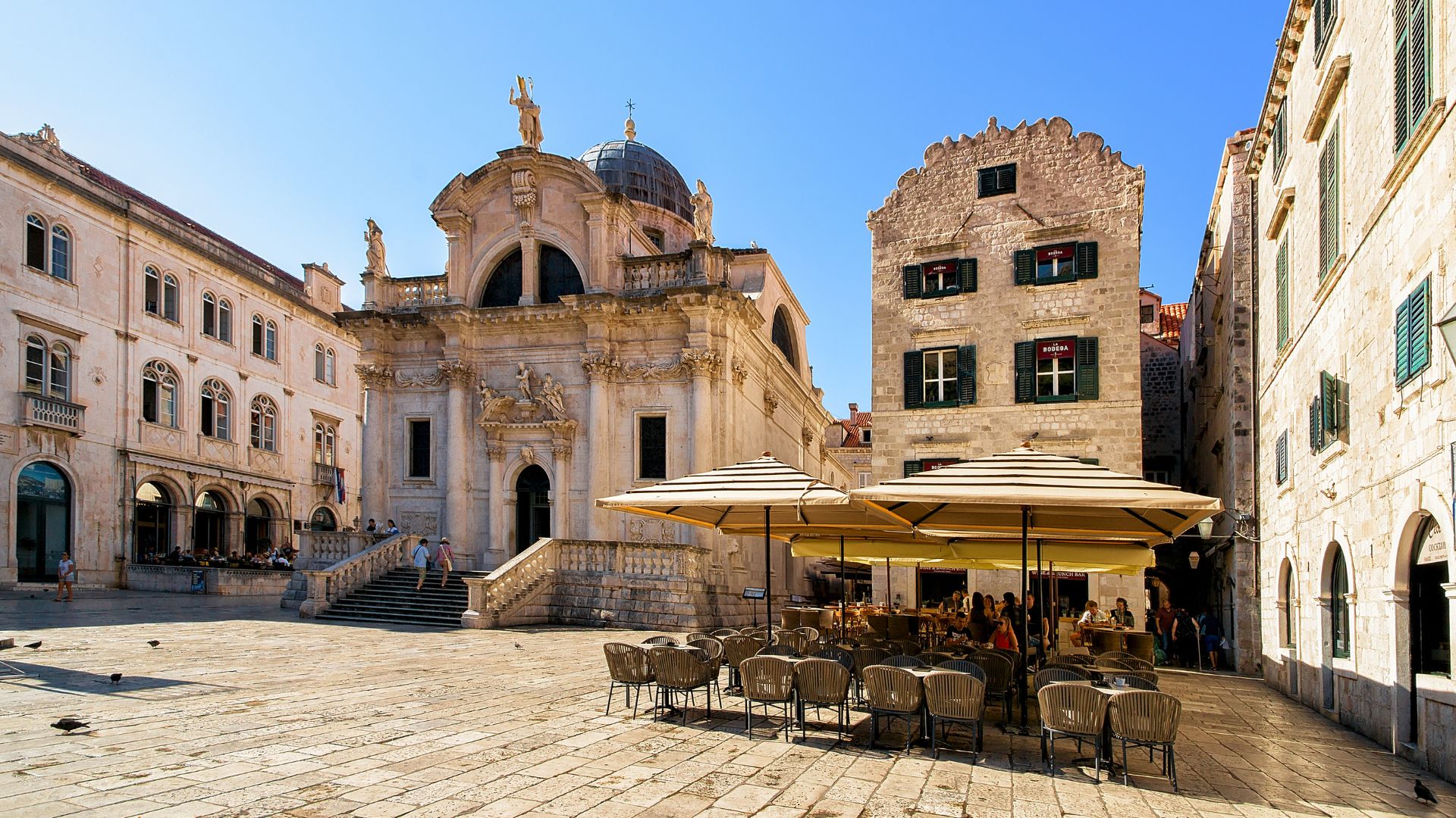 A wide-angle view of St. Blaise's Church, an 18th-century Baroque church, in the Old City of Dubrovnik, Croatia, on a sunny day. In the foreground, there's a paved square with outdoor seating and umbrellas from a cafe, while the grand church with its prominent dome and statues stands in the background, flanked by historic stone buildings. 
