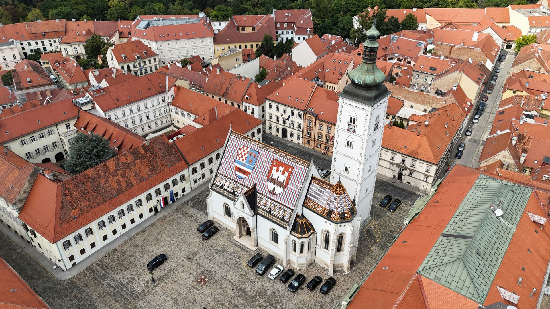 An aerial view of St. Mark's Church in Zagreb, Croatia, featuring its iconic colorful tiled roof depicting the coats of arms of Zagreb and the Triune Kingdom of Croatia, surrounded by traditional buildings with red-tiled roofs and a few cars parked in the square.