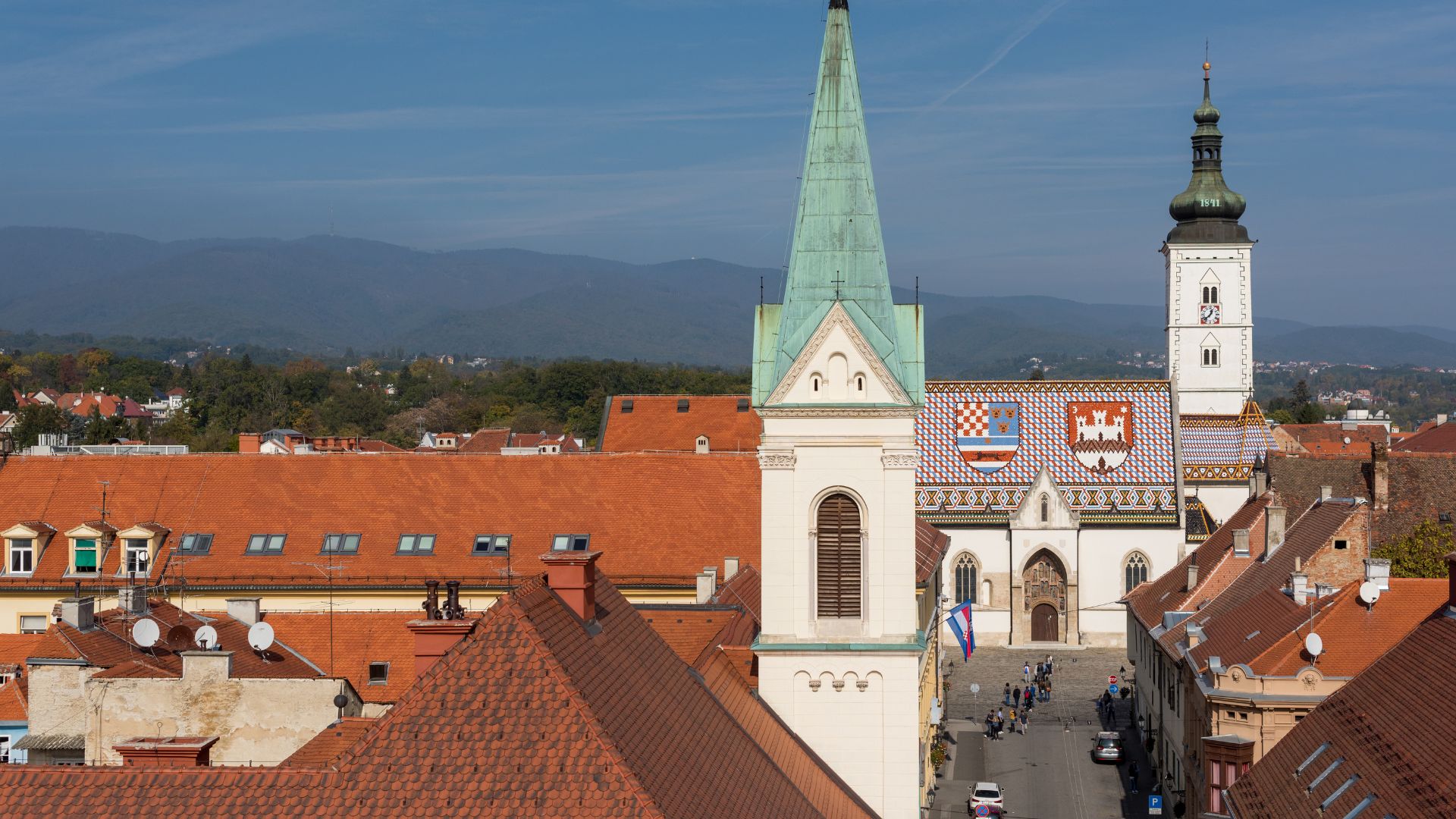 An aerial view of St. Mark's Church in Zagreb, Croatia, featuring its distinctive colorful tiled roof depicting the coats of arms of Croatia, Dalmatia, Slavonia, and the city of Zagreb, surrounded by red-tiled rooftops of other buildings under a clear blue sky.