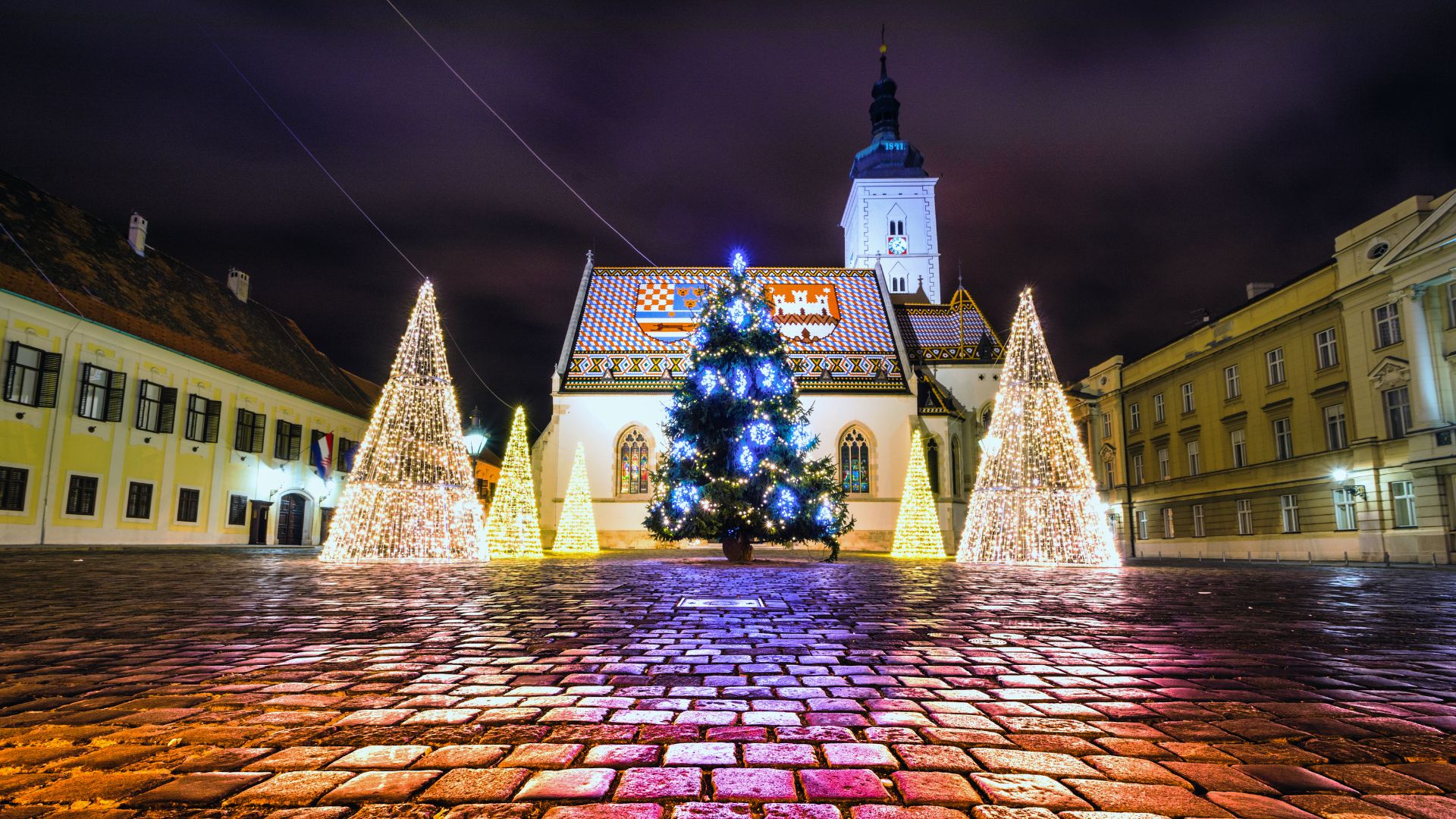 A night scene of St. Mark's Square in Zagreb, Croatia, during Advent, featuring St. Mark's Church adorned with Christmas lights and a large, brightly lit Christmas tree in the foreground, surrounded by several smaller illuminated conical trees.