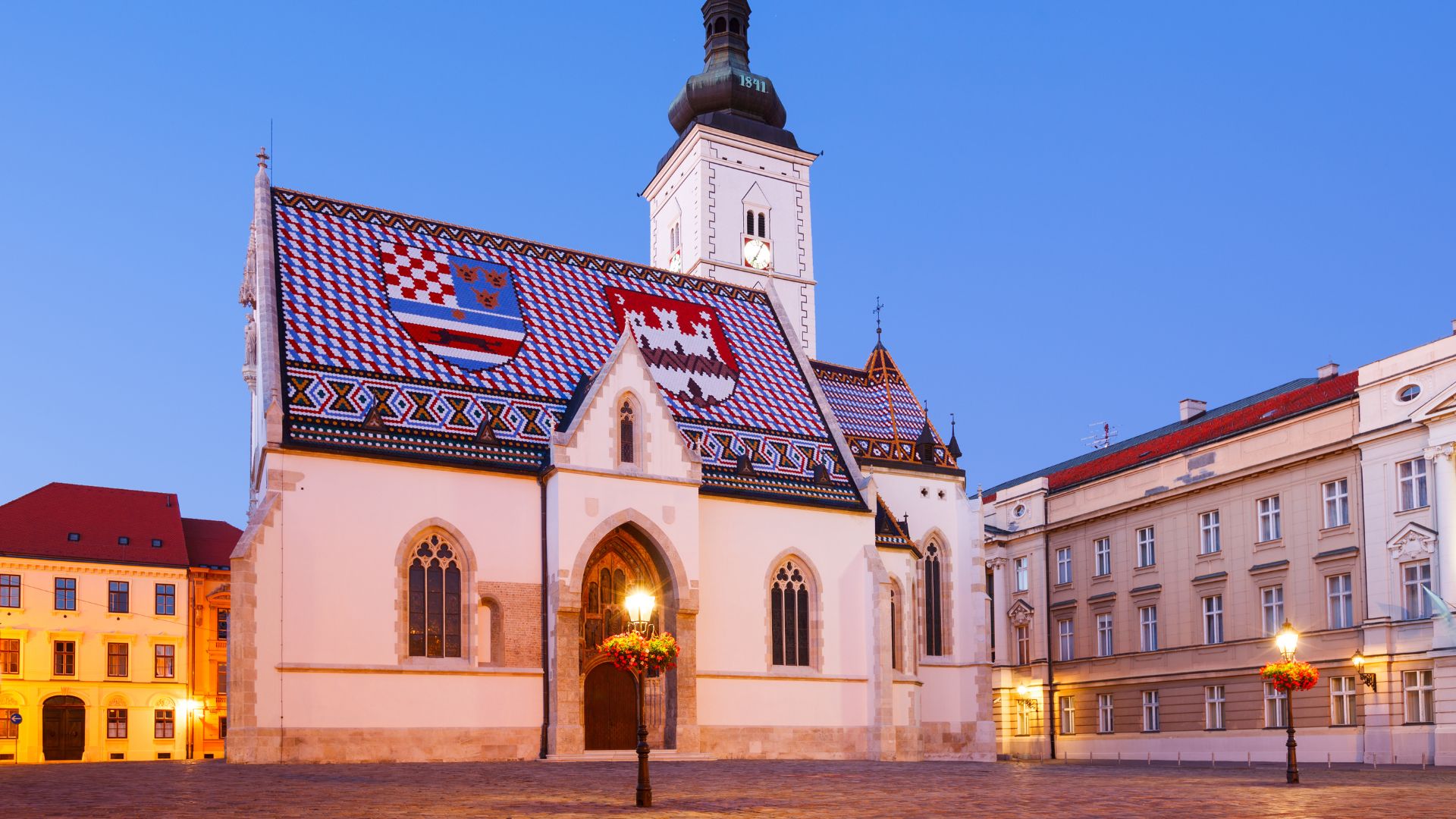 A low-angle, eye-level shot of St. Mark's Church in Zagreb's Upper Town under a clear blue sky at dusk, with its iconic colorful tiled roof featuring the coats of arms of Croatia and Zagreb, and a tall white bell tower rising behind it. The church is illuminated by warm streetlights, and historical buildings flank the square on either side.