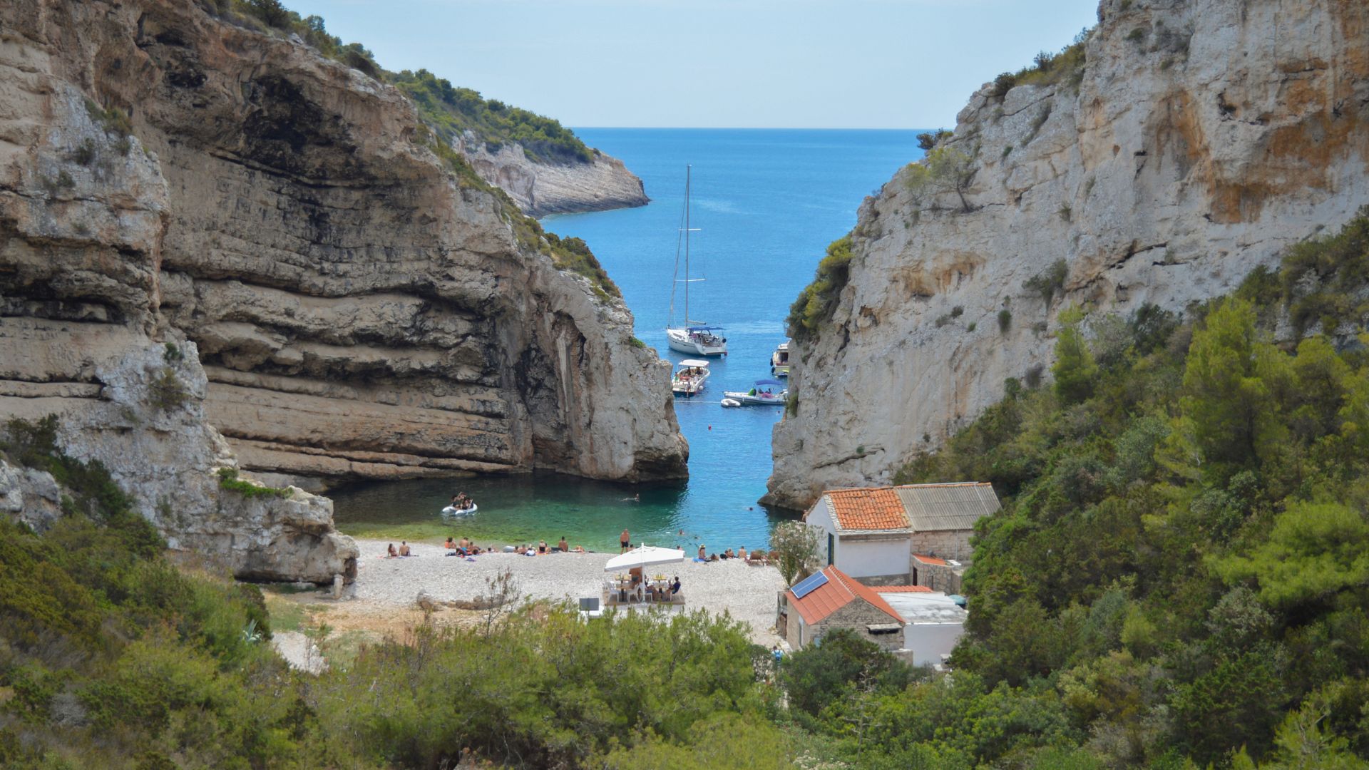 A scenic view of Stiniva Beach in Croatia, showing a secluded pebble beach and turquoise water nestled between towering cliffs, with boats anchored in the cove and a few buildings visible near the shore.