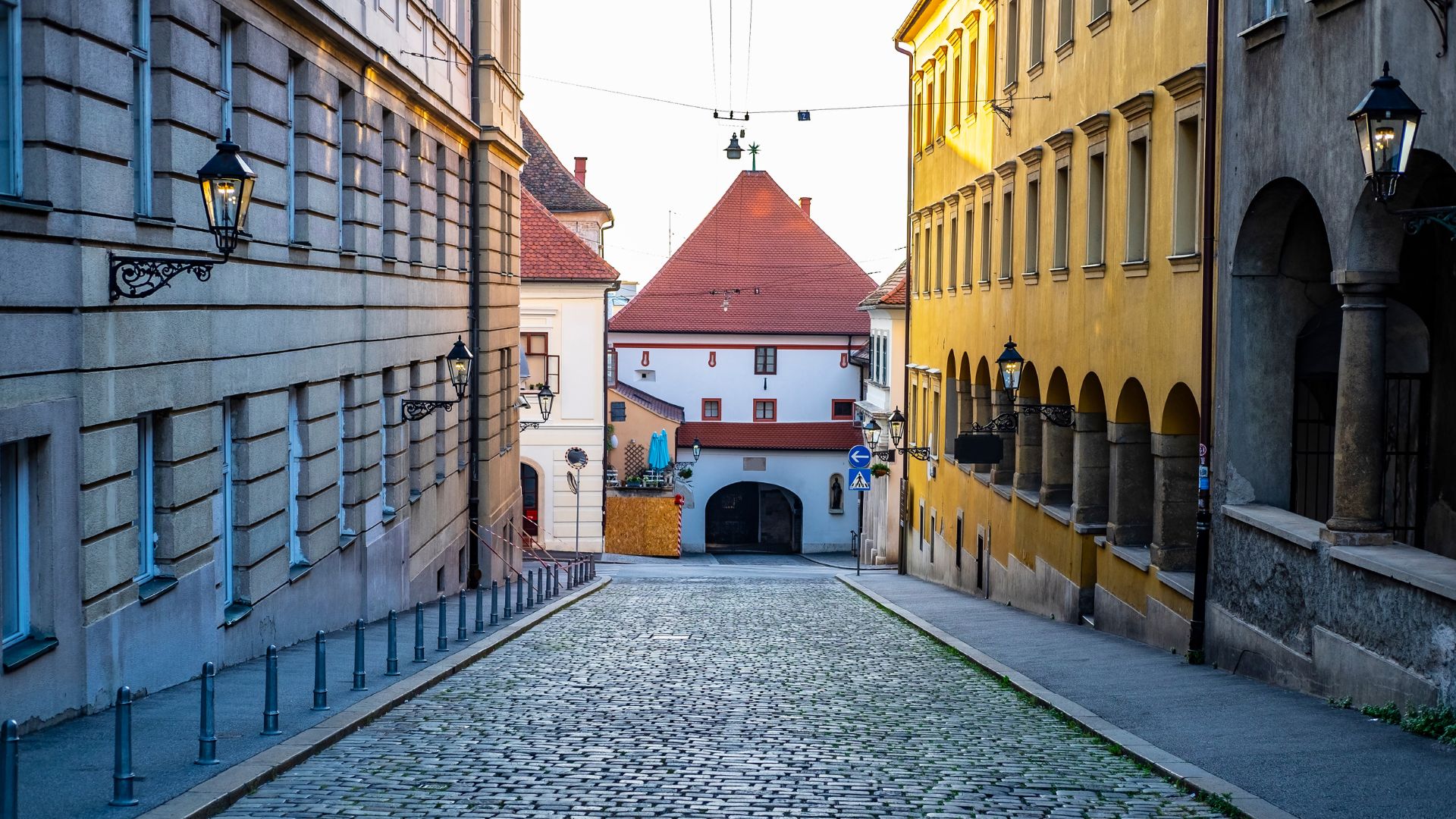 A cobblestone street in Zagreb's Upper Town leads towards the historic Stone Gate, flanked by old buildings with traditional architecture and street lamps.