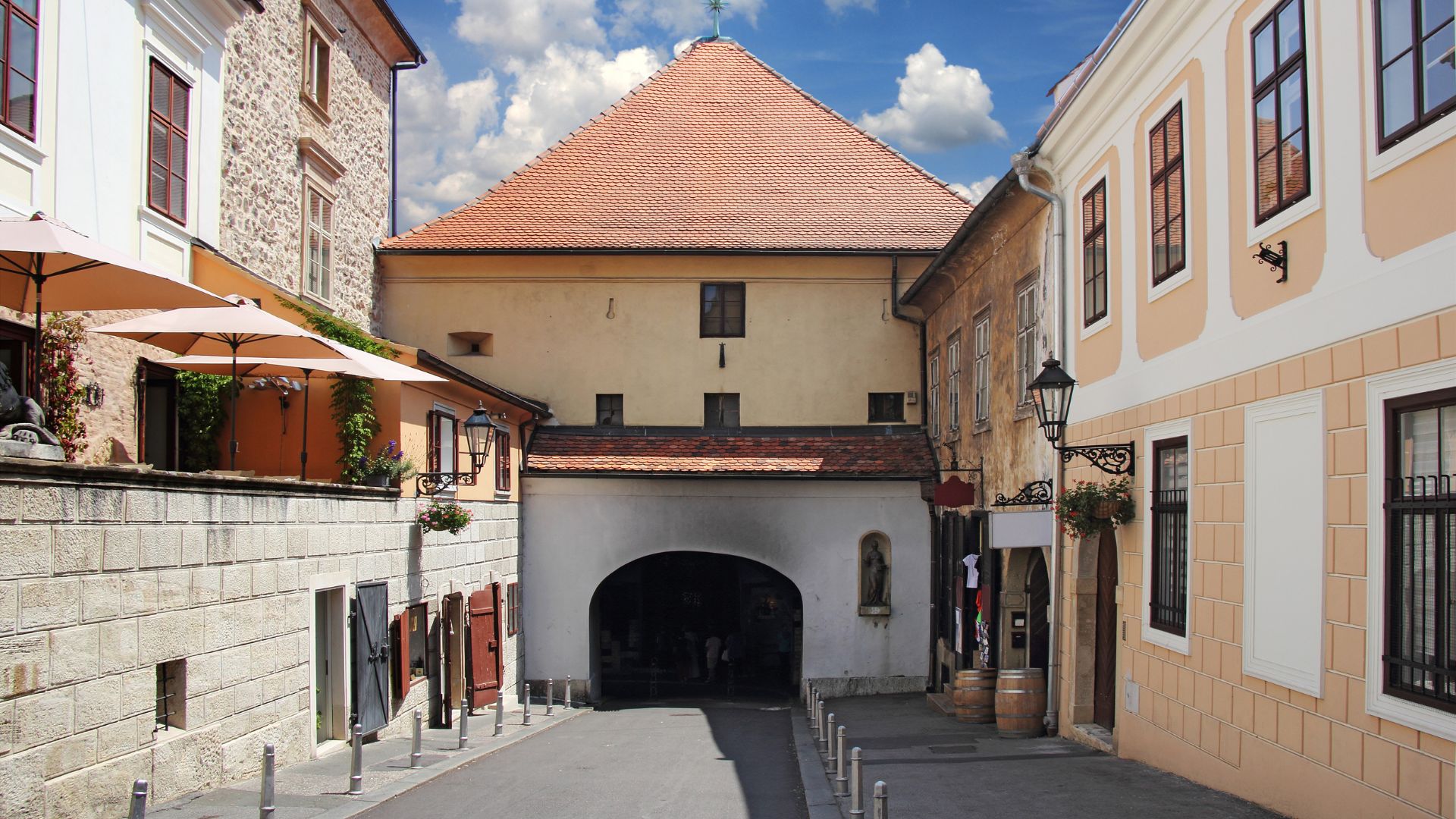 A narrow, cobblestone street in an old European town leads to an arched stone gate with a red-tiled roof structure above it, flanked by multi-story buildings on either side under a blue sky with scattered clouds.