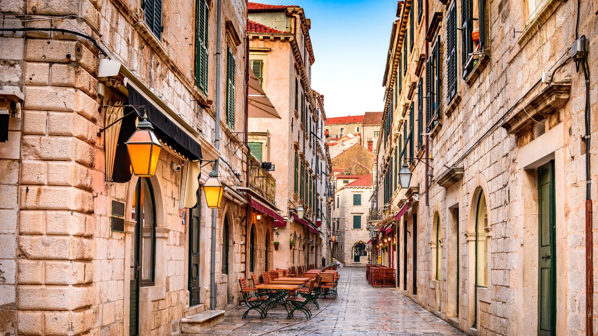 A narrow, paved street lined with historic stone buildings, featuring outdoor seating areas for cafes or restaurants on one side and old-fashioned lanterns illuminating the path. The street recedes into the distance, revealing more of the ancient city's architecture under a clear sky.