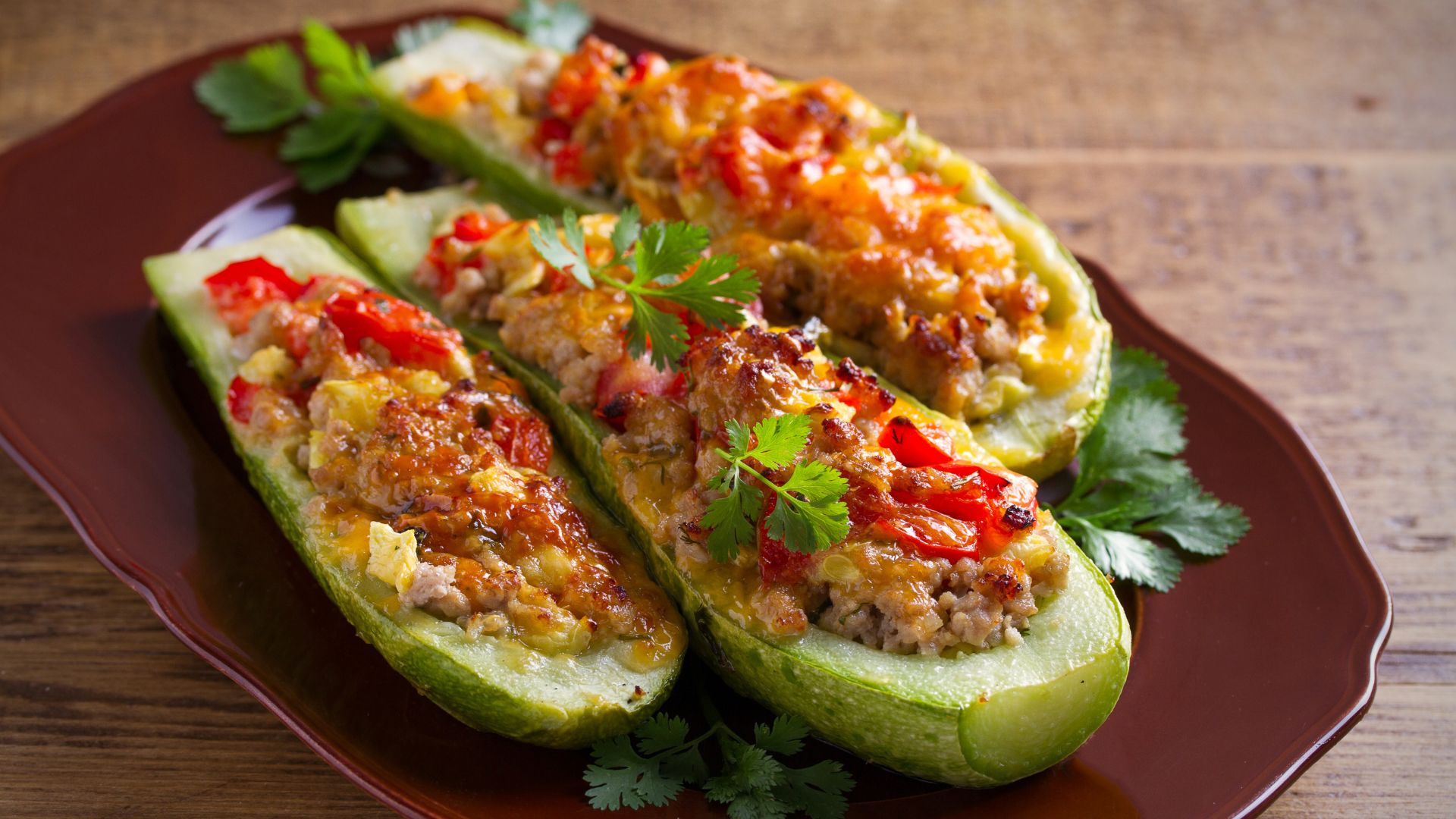 A close-up shot of three stuffed zucchini halves (punjene tikvice) arranged on a brown plate. The zucchini boats are filled with a mixture of ground meat, rice, and tomatoes, topped with melted cheese and garnished with fresh parsley. The dish is presented on a wooden surface.