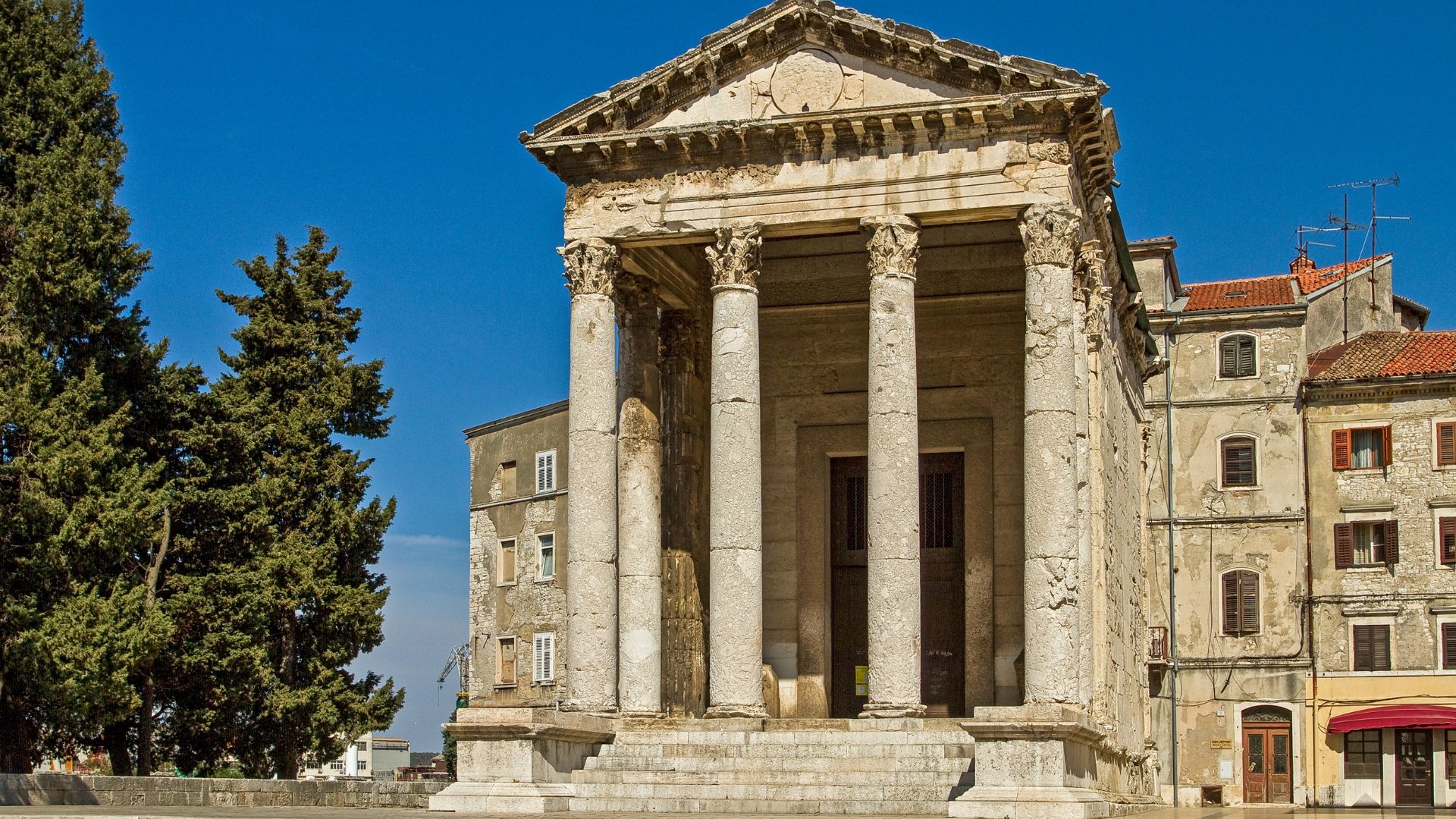 A well-preserved ancient Roman temple with a grand facade featuring four tall Corinthian columns and a triangular pediment, set against a clear blue sky, with older buildings visible in the background to the right and a large tree to the left.