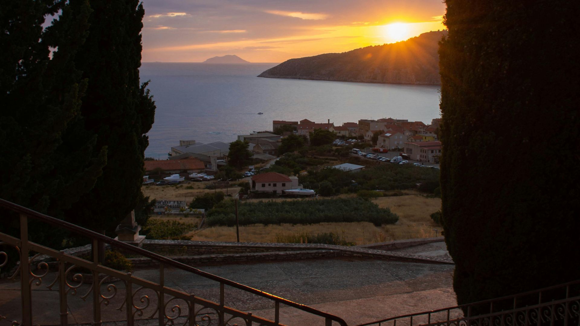 A sunset view over the coastal town of Komiža, Croatia, showing a bay with a distant island, framed by tall cypress trees and a stone staircase in the foreground.