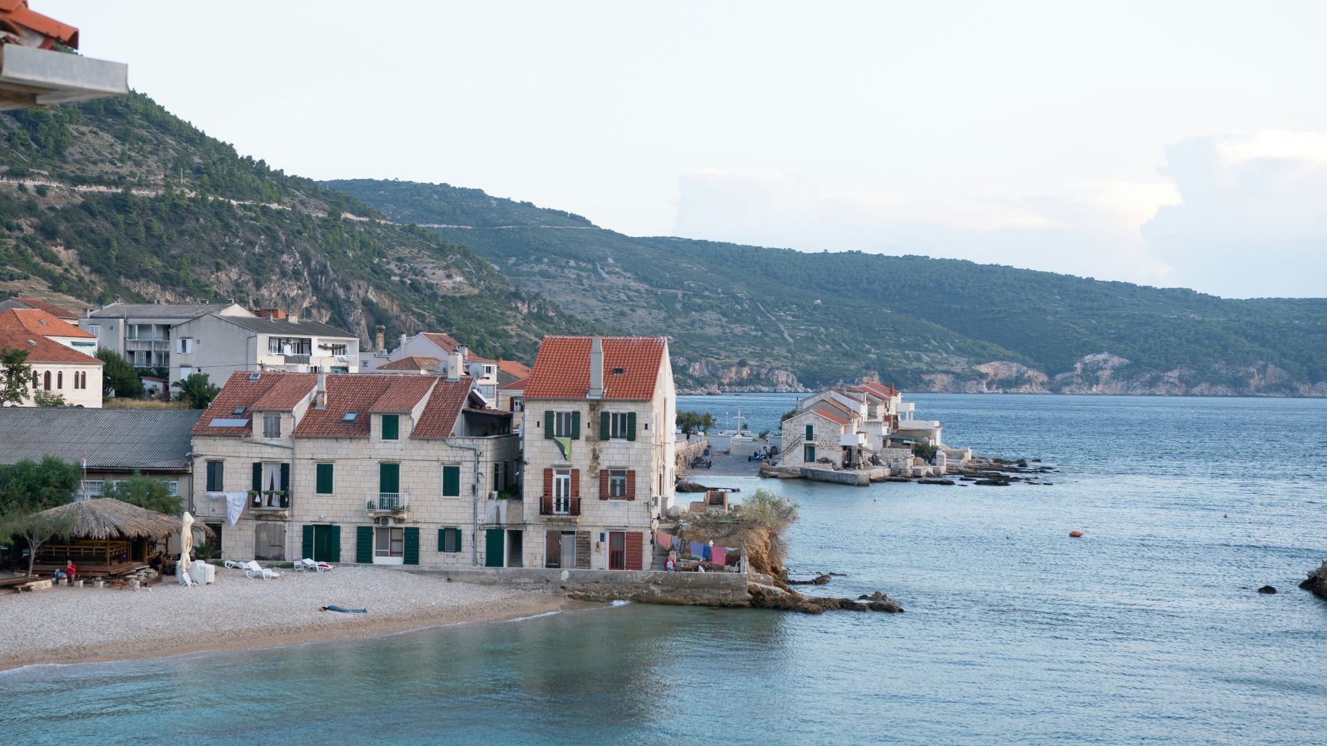 A scenic view of a coastal town with traditional stone houses featuring red-tiled roofs and green shutters, lining a pebble beach next to clear blue waters, with lush green hills rising in the background under a clear sky.
