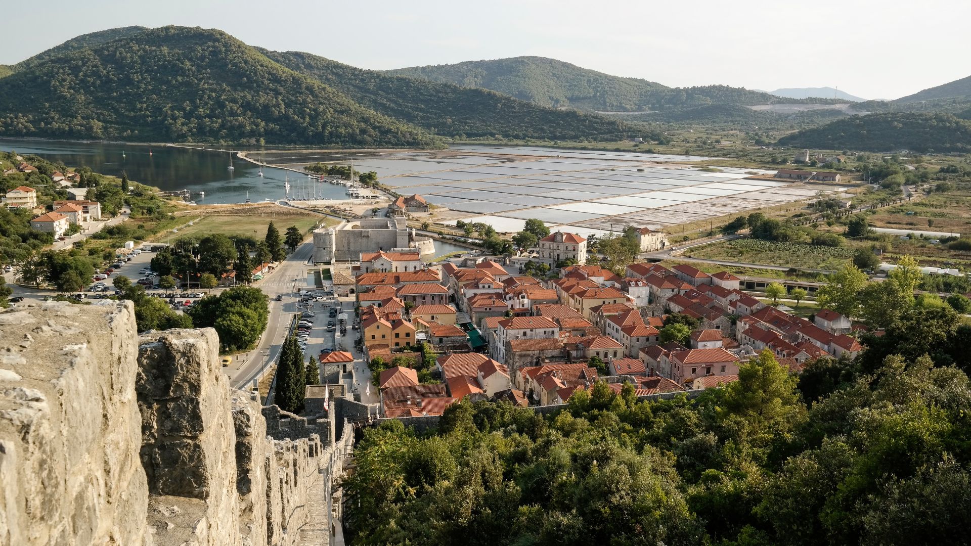An aerial view of the walled town of Ston, Croatia, nestled between hills and a body of water, with visible salt evaporation ponds and a long defensive wall snaking through the landscape.