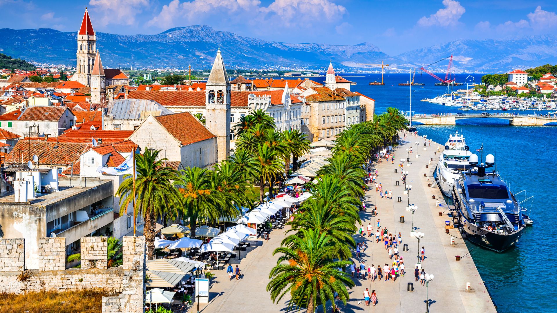 A vibrant aerial view of Trogir, Croatia, showcasing its historic red-tiled rooftops, a prominent bell tower, and a lively waterfront promenade lined with palm trees, umbrellas, and people, with boats docked in the clear blue water of the Adriatic Sea and mountains in the background. 
