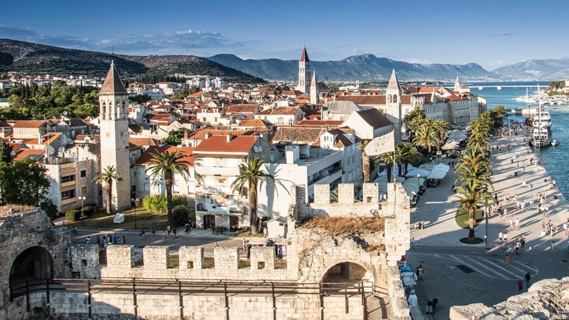A panoramic view of the historic city of Trogir, Croatia, showing its red-tiled roofs, ancient stone buildings, and prominent bell towers nestled alongside a calm blue waterway with boats, all set against a backdrop of distant mountains and a clear sky. 