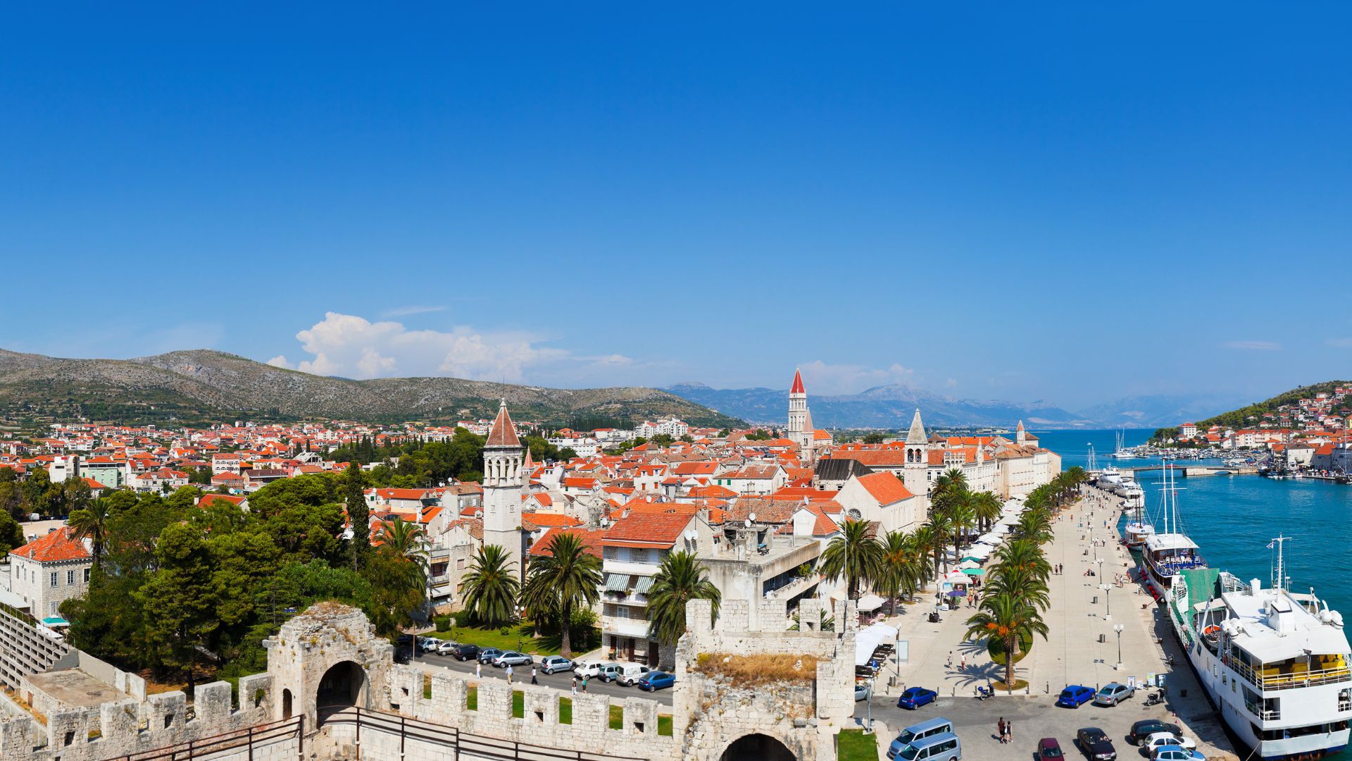 A panoramic view of the historic city of Trogir, Croatia, showing its red-tiled roofs, ancient stone buildings, and prominent bell towers nestled alongside a calm blue waterway with boats, all set against a backdrop of distant mountains and a clear sky.