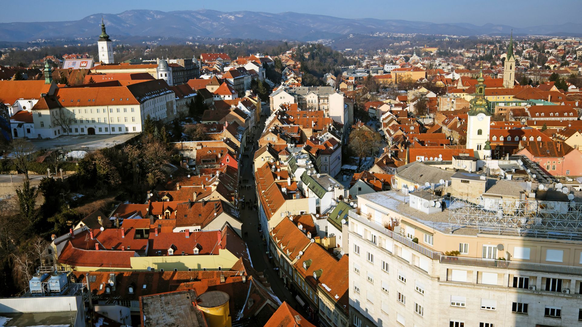 An aerial view of the historic Upper Town of Zagreb, Croatia, featuring a dense arrangement of buildings with orange-tiled roofs, church steeples, and winding streets, set against a backdrop of distant mountains.