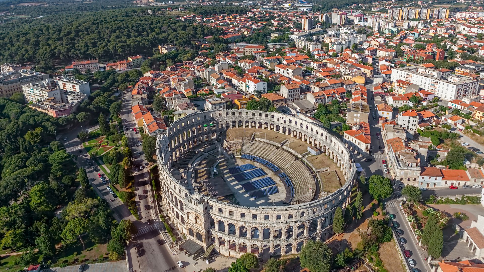 An aerial view of the well-preserved Pula Arena, a large oval Roman amphitheater, surrounded by a densely built city and green trees.