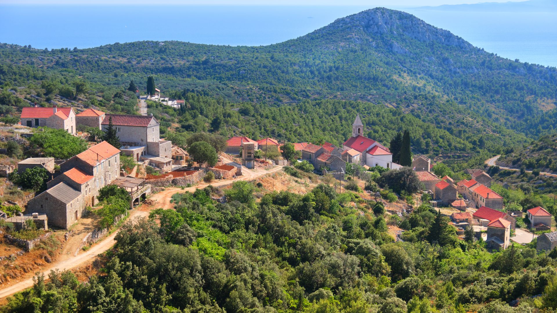 An aerial view shows the village of Velo Grablje on Hvar Island, Croatia, with stone houses and a church nestled on a hillside covered in lush green vegetation, overlooking the sea in the distance.