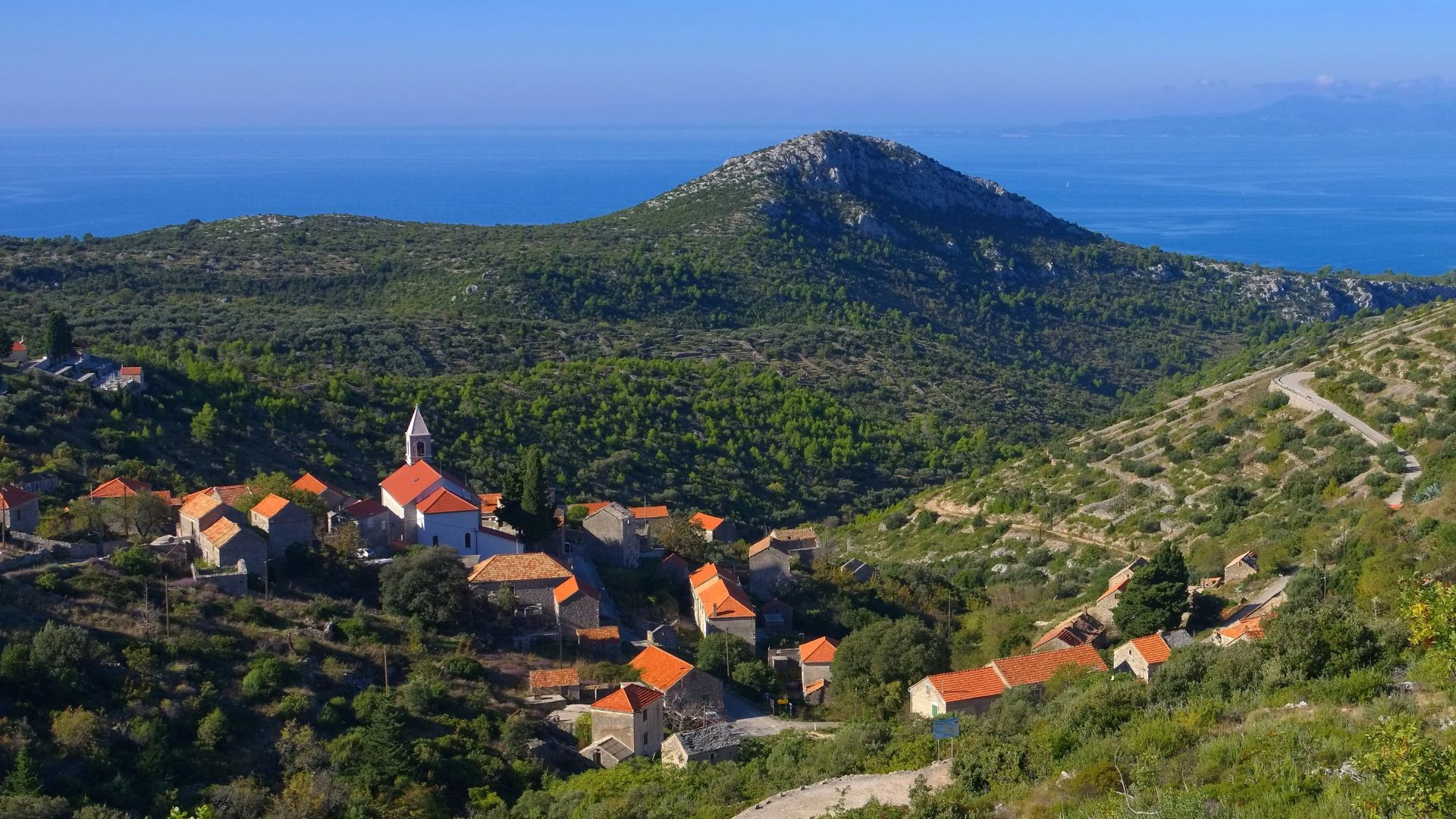 A picturesque village with terracotta-roofed stone houses and a prominent church tower nestled in a green valley, surrounded by hills and with the blue sea and distant islands visible in the background under a clear sky.