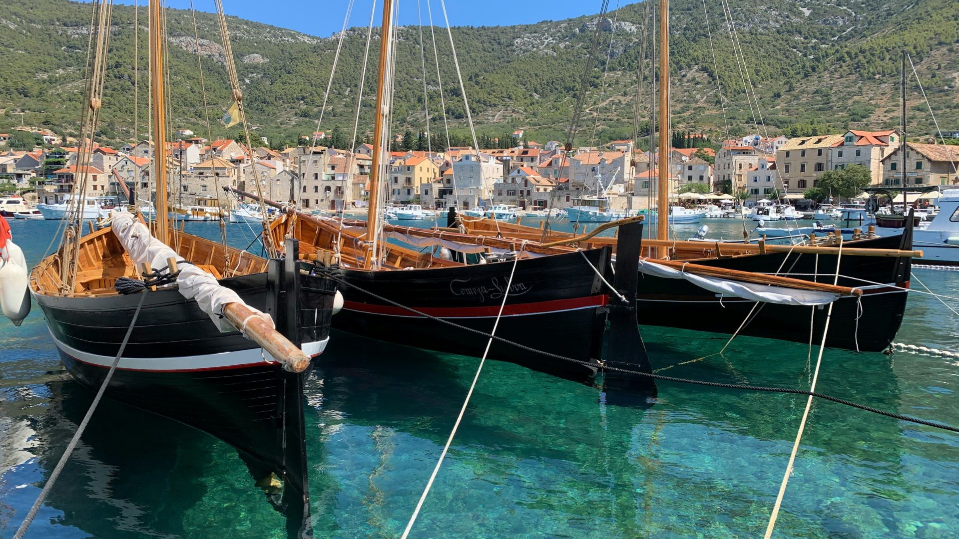 The image shows three traditional wooden sailing boats with black hulls and wooden masts docked in a clear, turquoise harbor. In the background, a picturesque coastal town with terracotta-roofed buildings nestles at the foot of a green, mountainous landscape under a bright blue sky. 