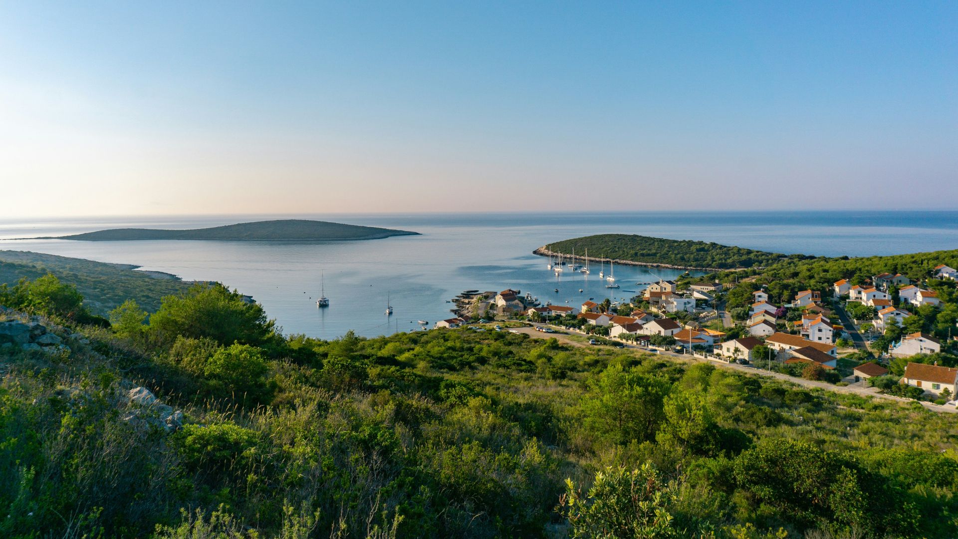 A high-angle view of a coastal town with red-roofed houses nestled among green hills, overlooking a calm bay with boats and two small, lush islands under a clear blue sky.