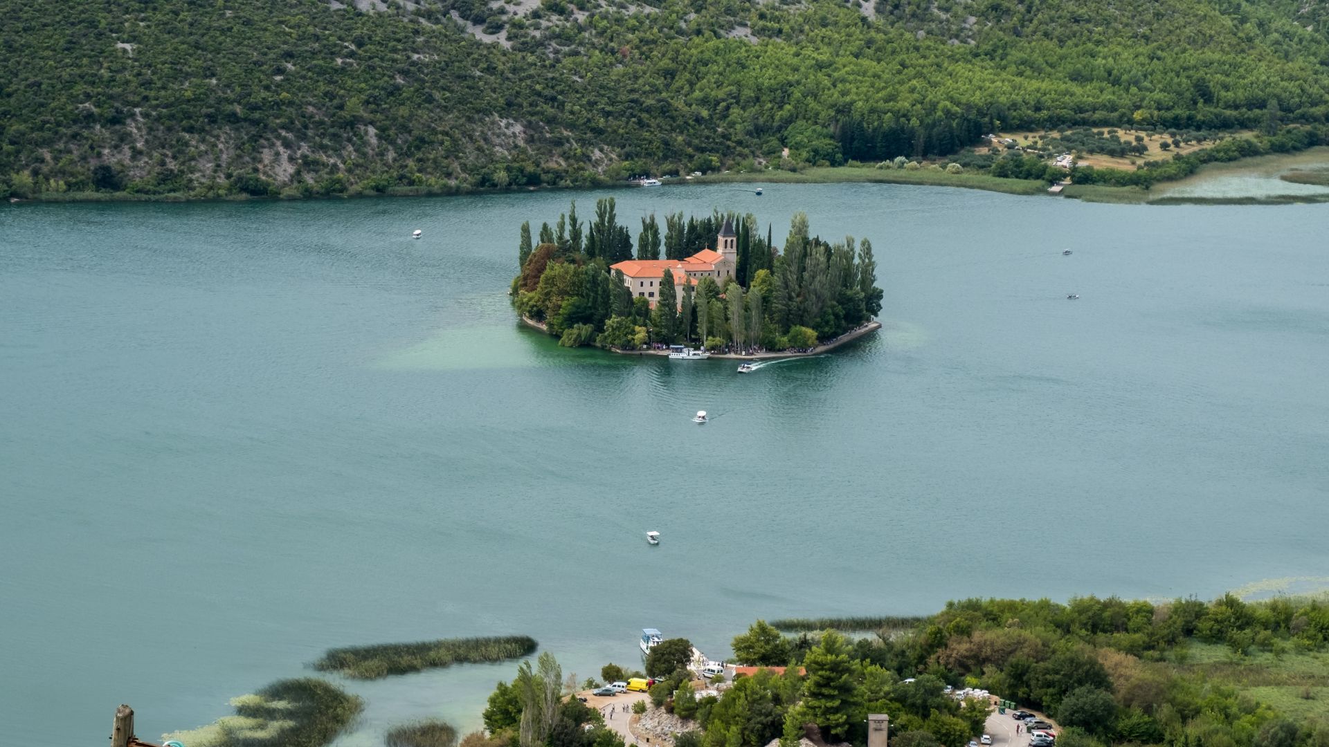 An aerial view of Visovac Monastery, a historic building complex surrounded by dense green trees on a small island in the calm, turquoise-green waters of Visovac Lake, with forested hills visible in the background.
