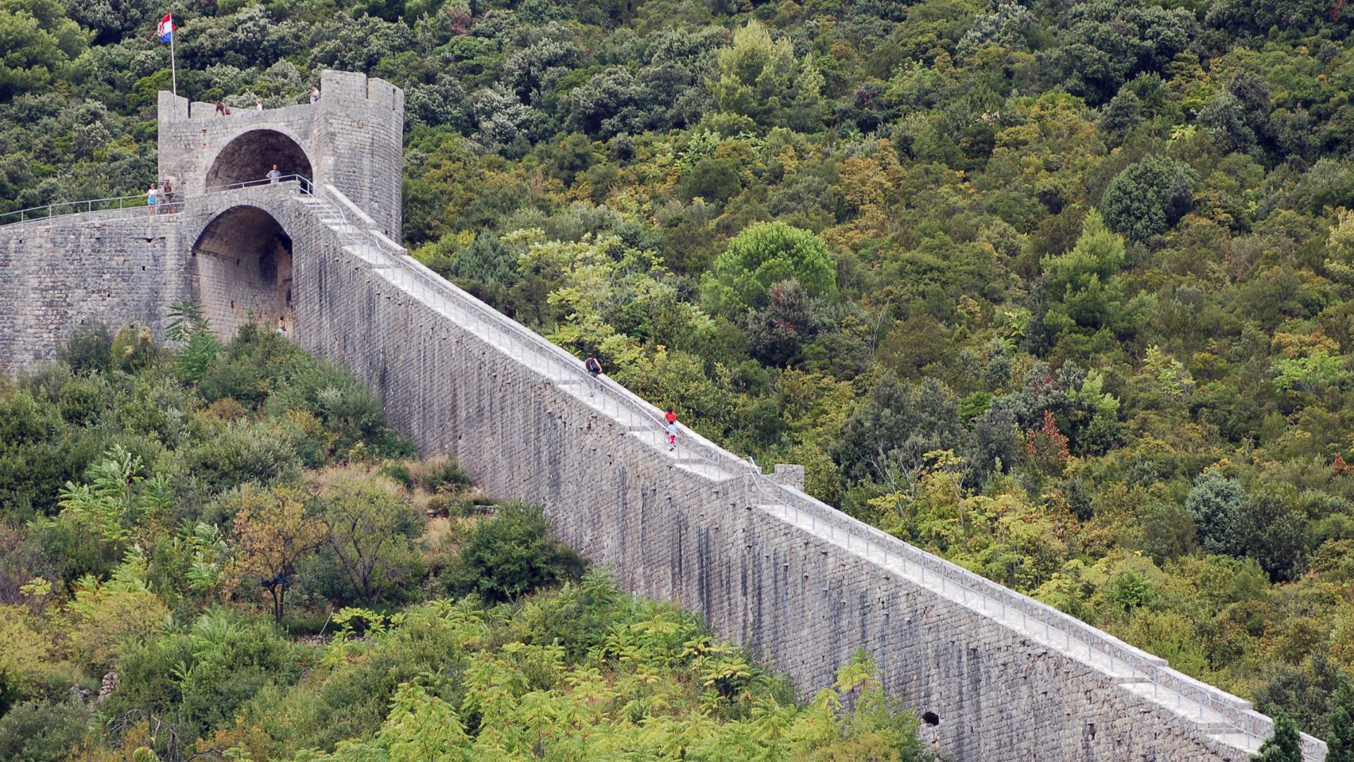A long, ancient stone wall with arches and a tower at one end stretches diagonally up a lush, green, forested hillside under a bright sky. People can be seen walking along the top of the wall.