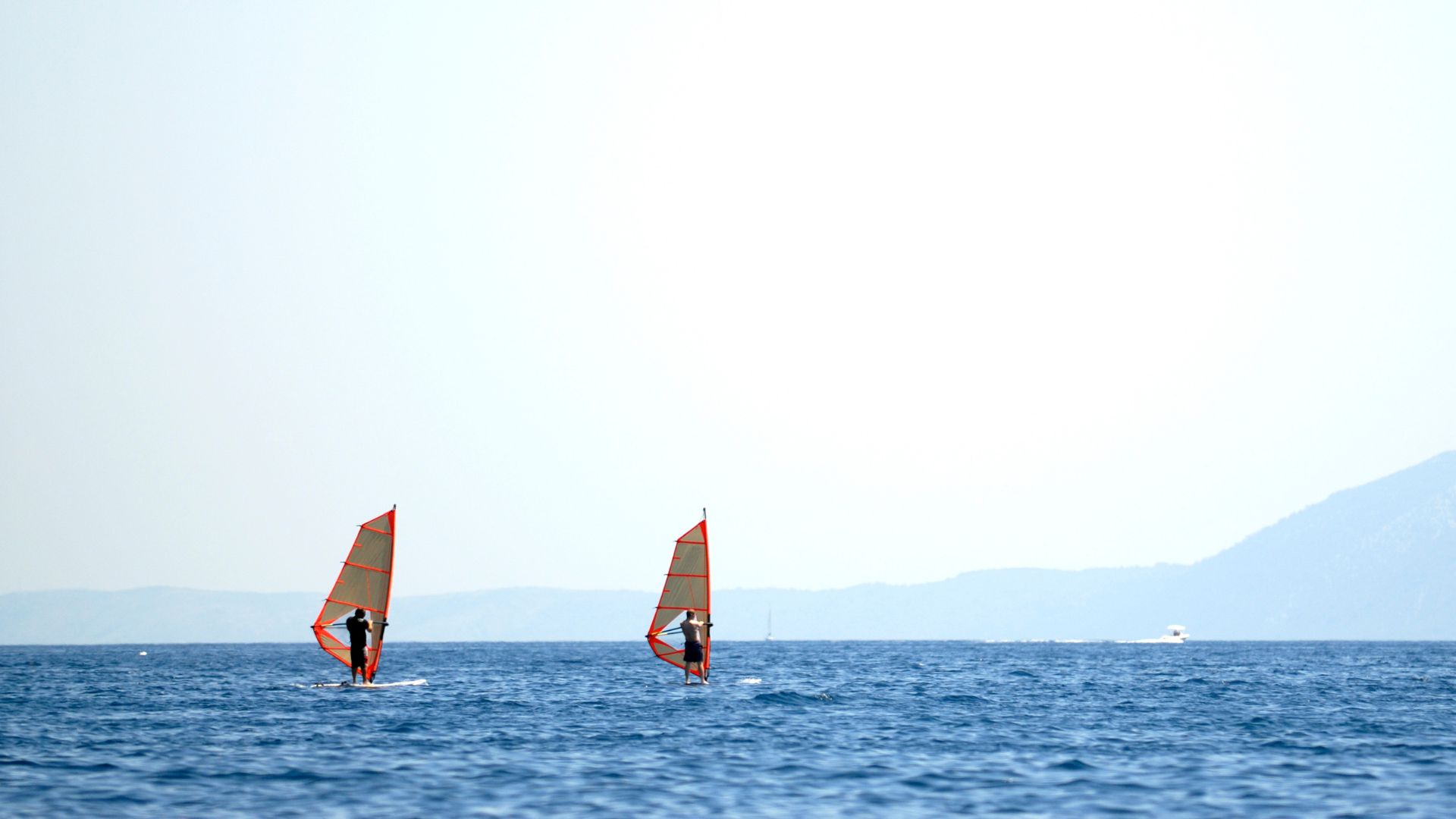 Two windsurfers are on a calm, blue sea under a clear sky, with a hazy mountain range visible in the distance.