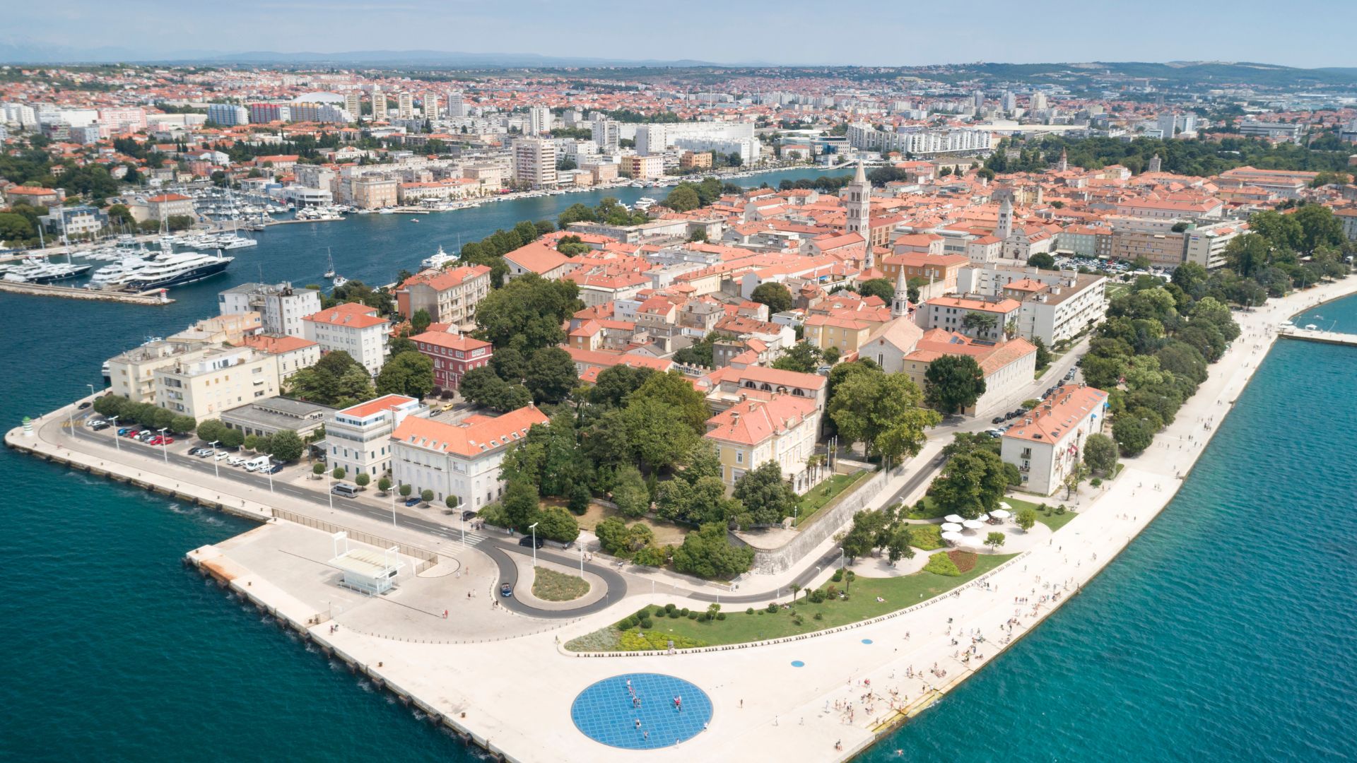 Aerial view of Zadar, Croatia, showing its distinctive peninsula with a historic cityscape, surrounded by the clear blue waters of the Adriatic Sea, with boats in the harbor and modern structures along the waterfront.