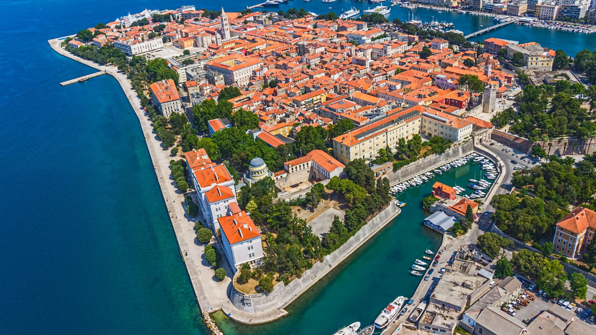 Aerial view of Zadar, Croatia, a coastal city with a densely packed historic center featuring red-tiled roofs, surrounded by the clear blue waters of the Adriatic Sea and a marina filled with boats.