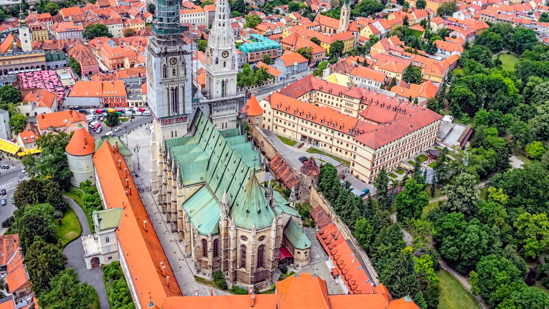 An aerial view of Zagreb Cathedral with its tall spires and green roof, surrounded by other buildings with orange-tiled roofs, and lush green trees in Zagreb's historic Upper Town.