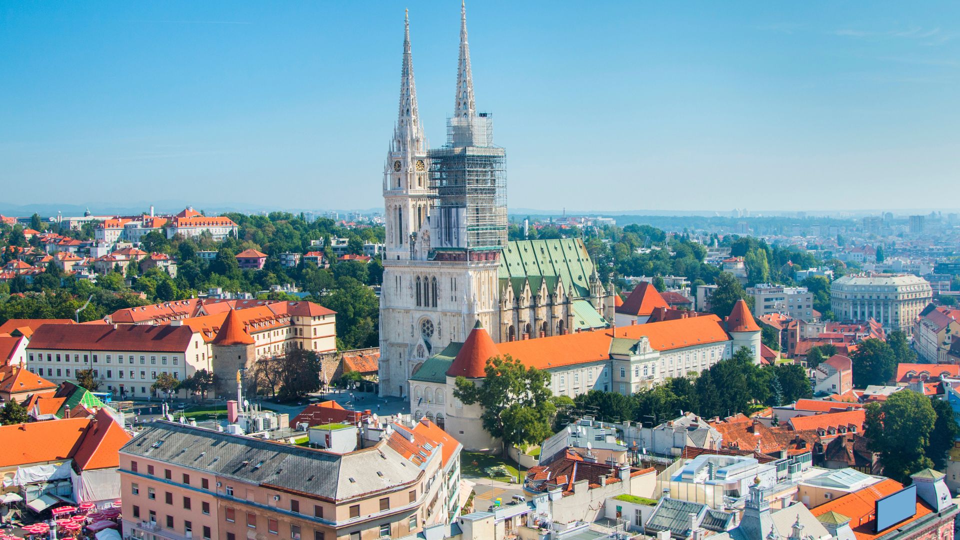 An aerial view of the Zagreb Cathedral, a grand Neo-Gothic structure with two prominent spires, rising above the surrounding cityscape of red-roofed buildings and lush green trees under a clear blue sky.