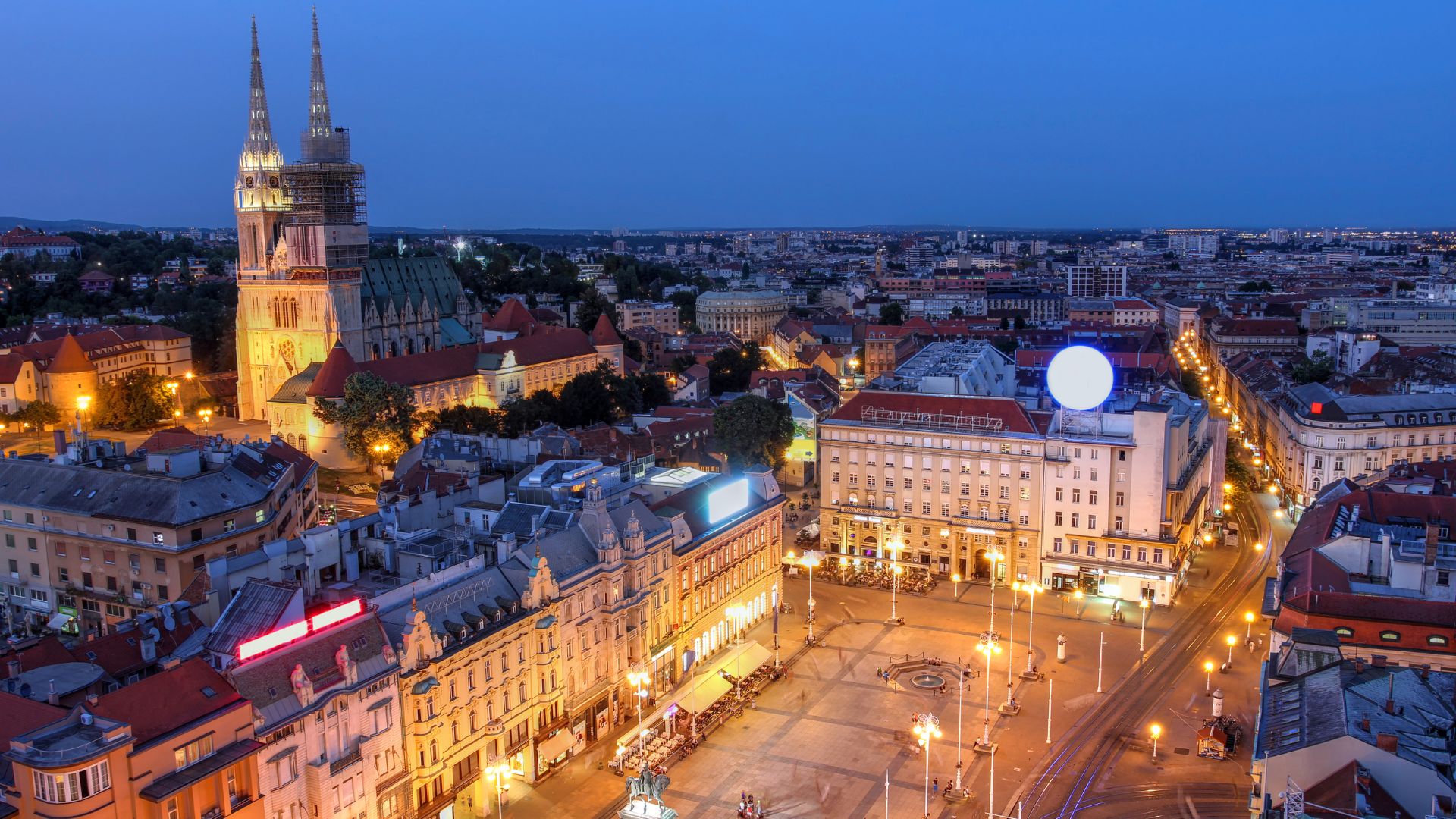 A high-angle nighttime view of a bustling European city square and surrounding buildings, illuminated by streetlights and building lights, with a large cathedral featuring prominent spires visible in the background against a dark blue sky.
