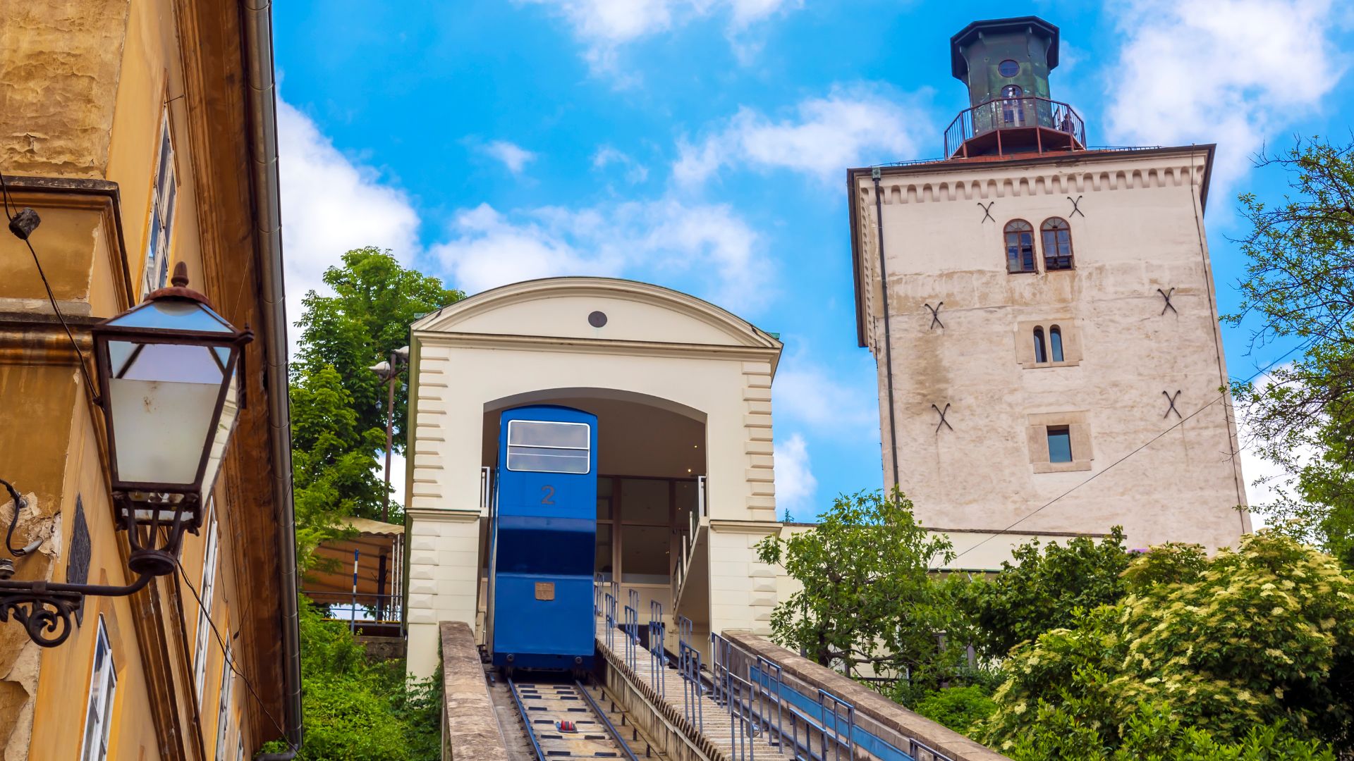 A blue funicular car ascends a track towards a white building with an arched entrance, situated next to the tall, light-colored Lotrščak Tower under a blue sky with white clouds.