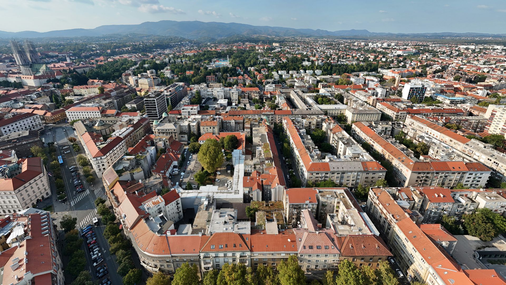 An aerial view captures the sprawling city of Zagreb, Croatia, with numerous buildings featuring red-tiled roofs, streets, and green spaces, all set against a backdrop of distant mountains under a clear blue sky.