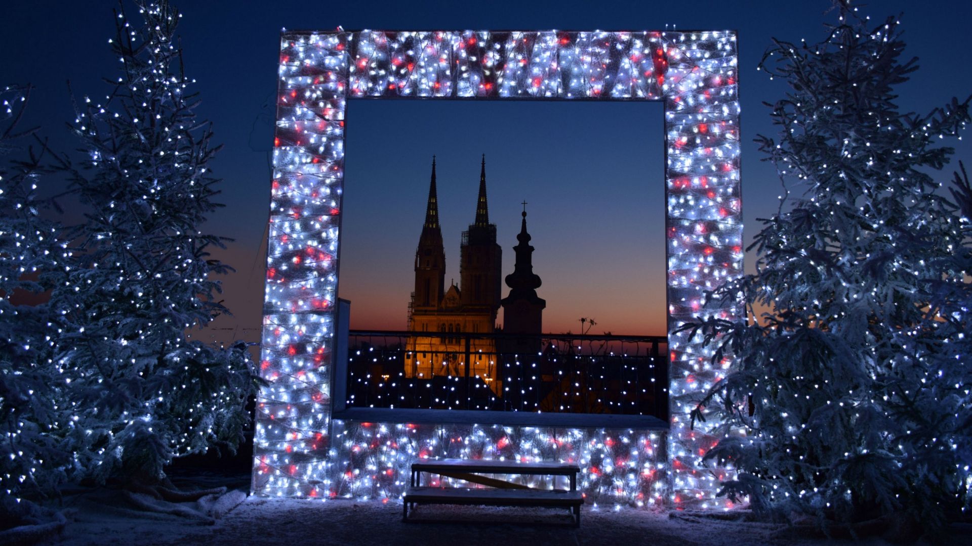 A nighttime view of the Zagreb Cathedral's spires, framed by a large, rectangular archway adorned with festive white and red lights, flanked by two snow-dusted Christmas trees also decorated with lights, with a small bench in the foreground.