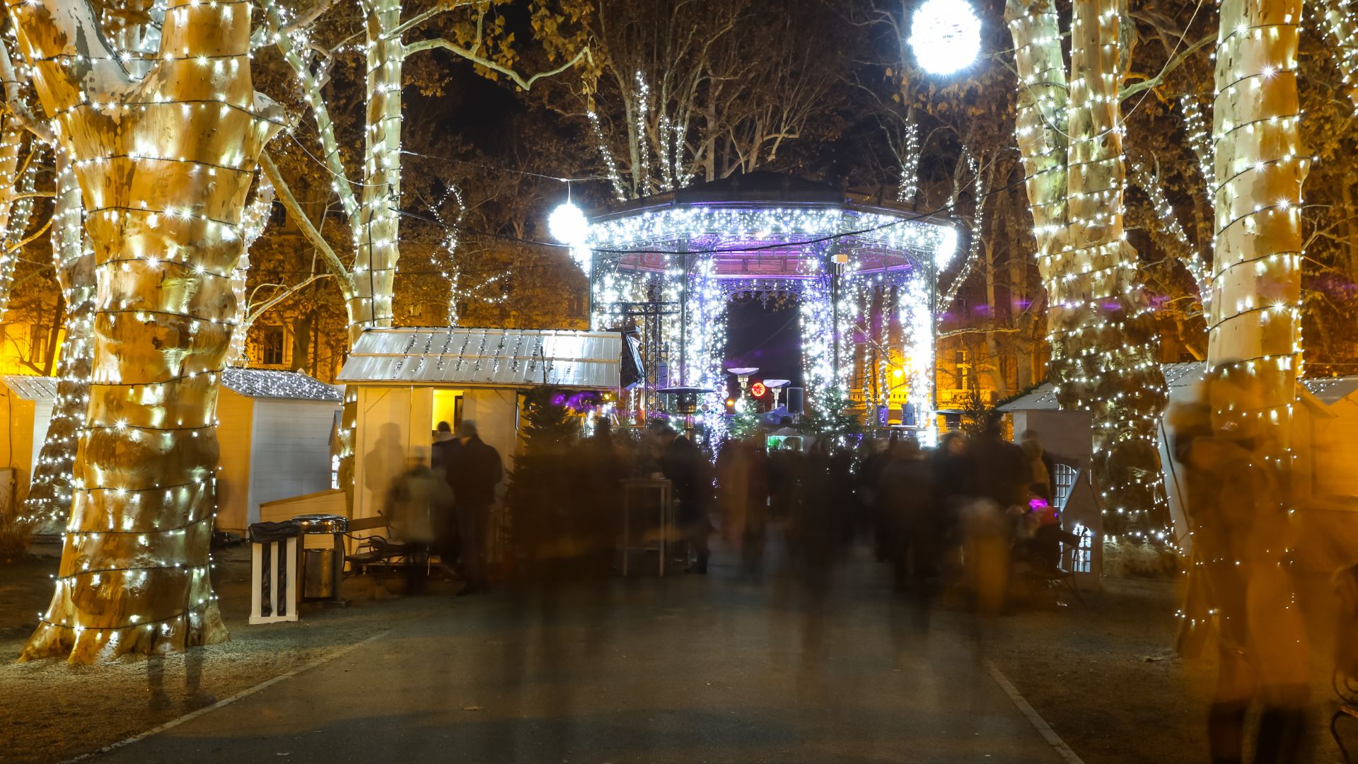 A nighttime scene of Zagreb's Advent market in Zrinjevac Park, featuring a brightly lit gazebo and trees wrapped in festive lights, with blurred figures of people moving through the illuminated space.