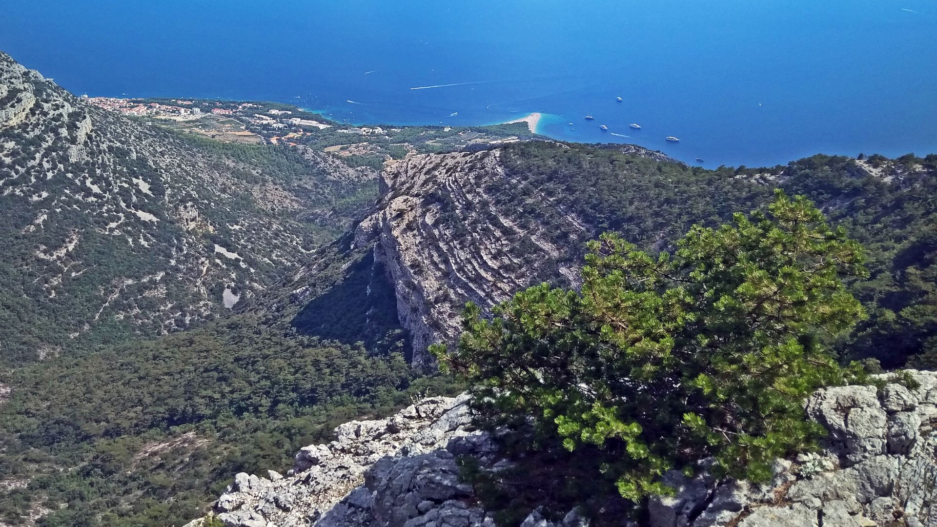 An aerial view from a mountain overlooking a clear blue sea with a distinctive, horn-shaped pebble beach (Zlatni Rat) extending into the water, surrounded by green hills and a distant coastline.