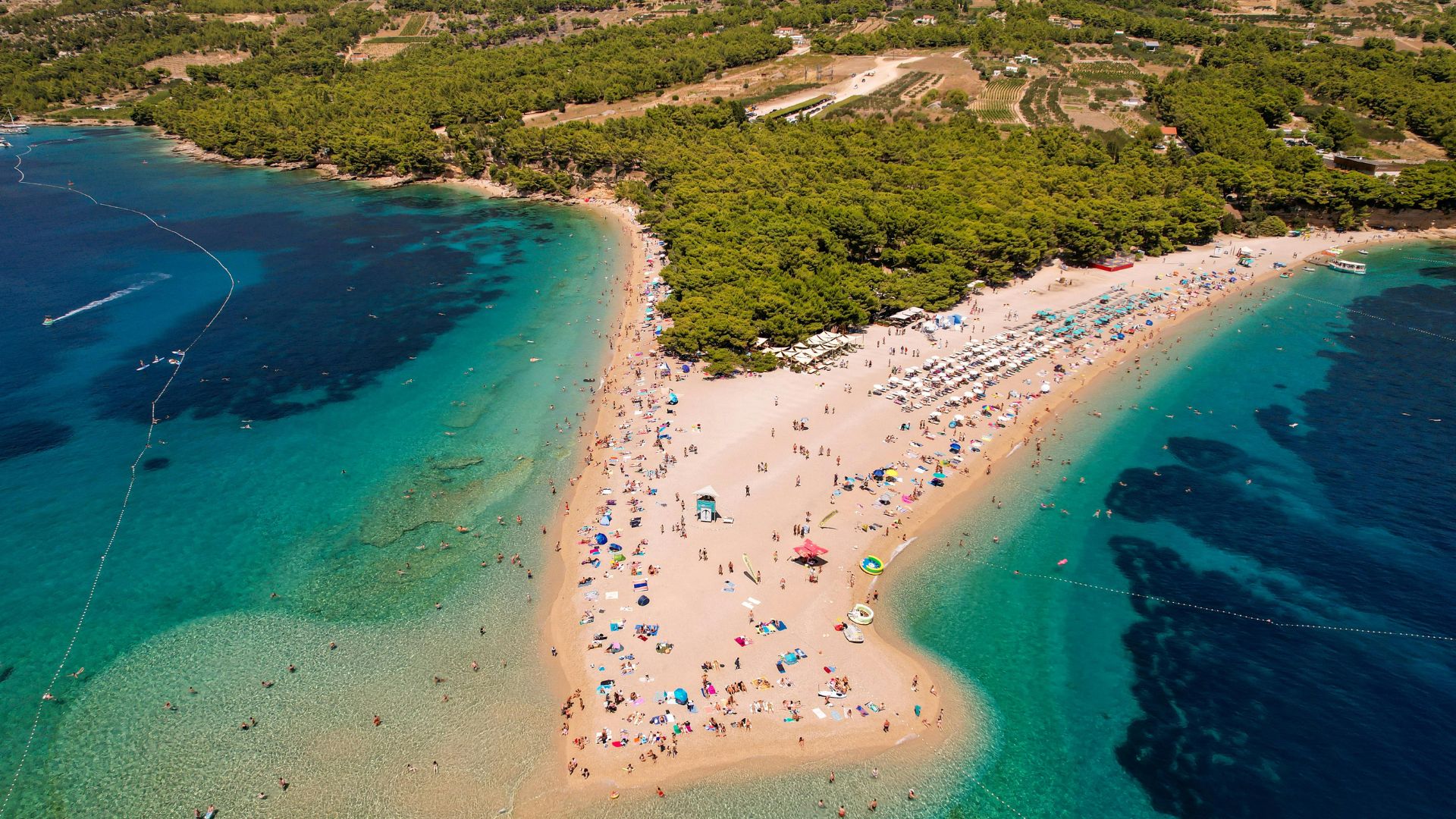 An aerial view shows the unique, V-shaped Zlatni Rat beach on Brač Island, Croatia, extending into the turquoise Adriatic Sea, with a lush green forest at its base and numerous beachgoers enjoying the sun and water on the pebble shore. 