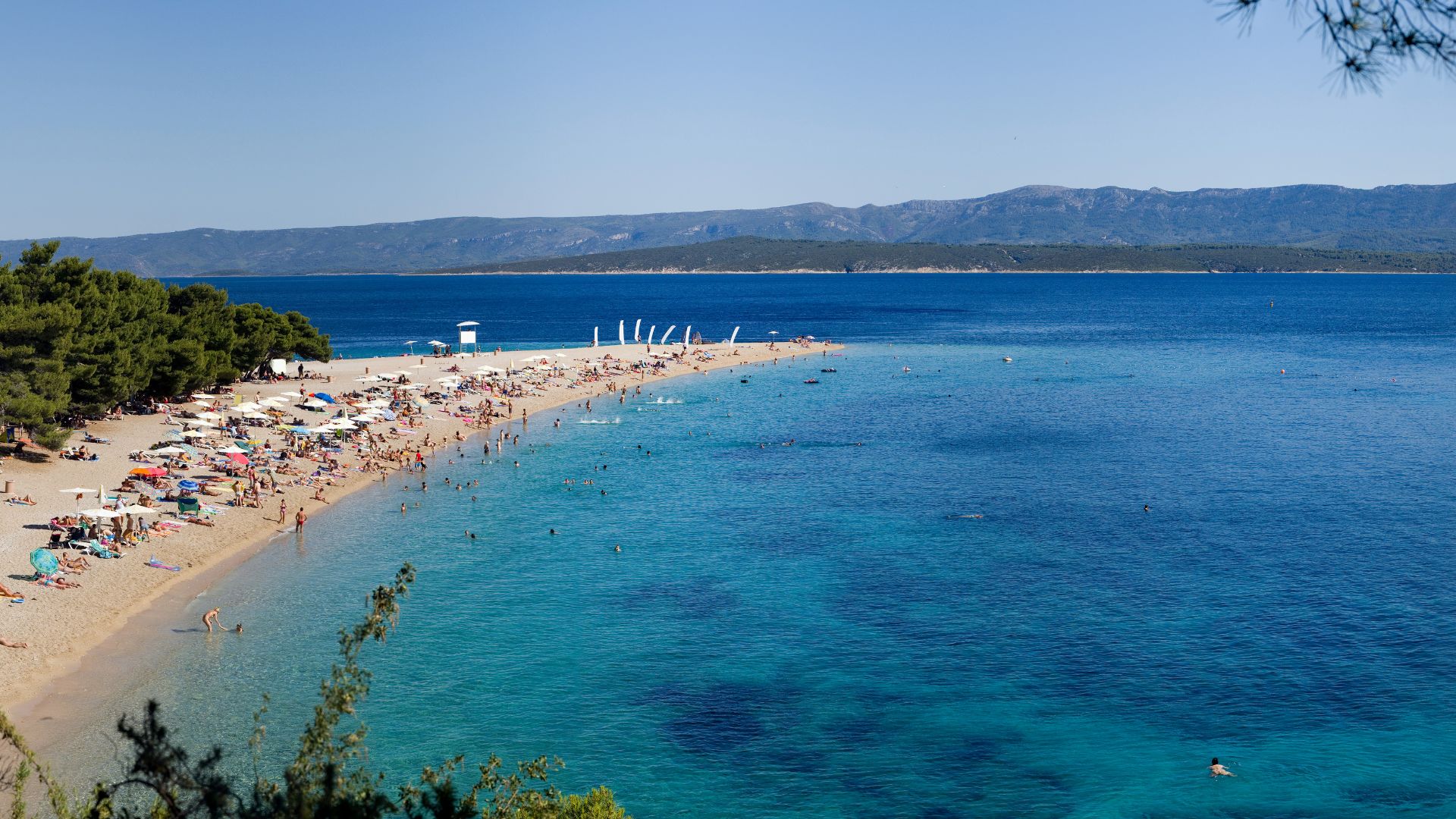 A scenic aerial view of Zlatni Rat beach (Golden Horn) on Brač Island, Croatia, showing its unique golden pebble spit extending into the clear turquoise waters of the Adriatic Sea, with numerous sunbathers and swimmers on the beach and in the water, and lush green trees lining the shore against a backdrop of distant mountains under a clear blue sky. 