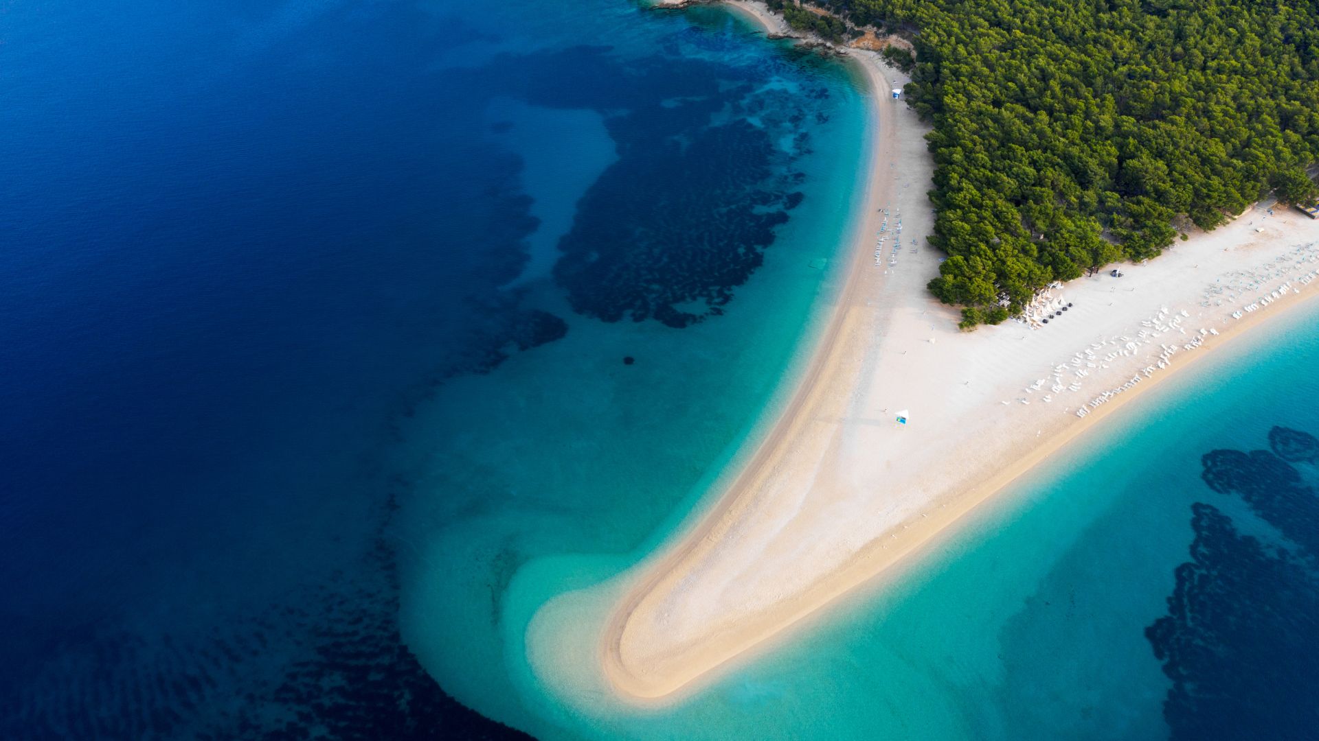 An aerial view of Zlatni Rat beach, a unique V-shaped pebble beach extending into the turquoise and deep blue waters of the Adriatic Sea, with a lush green forest at its base.