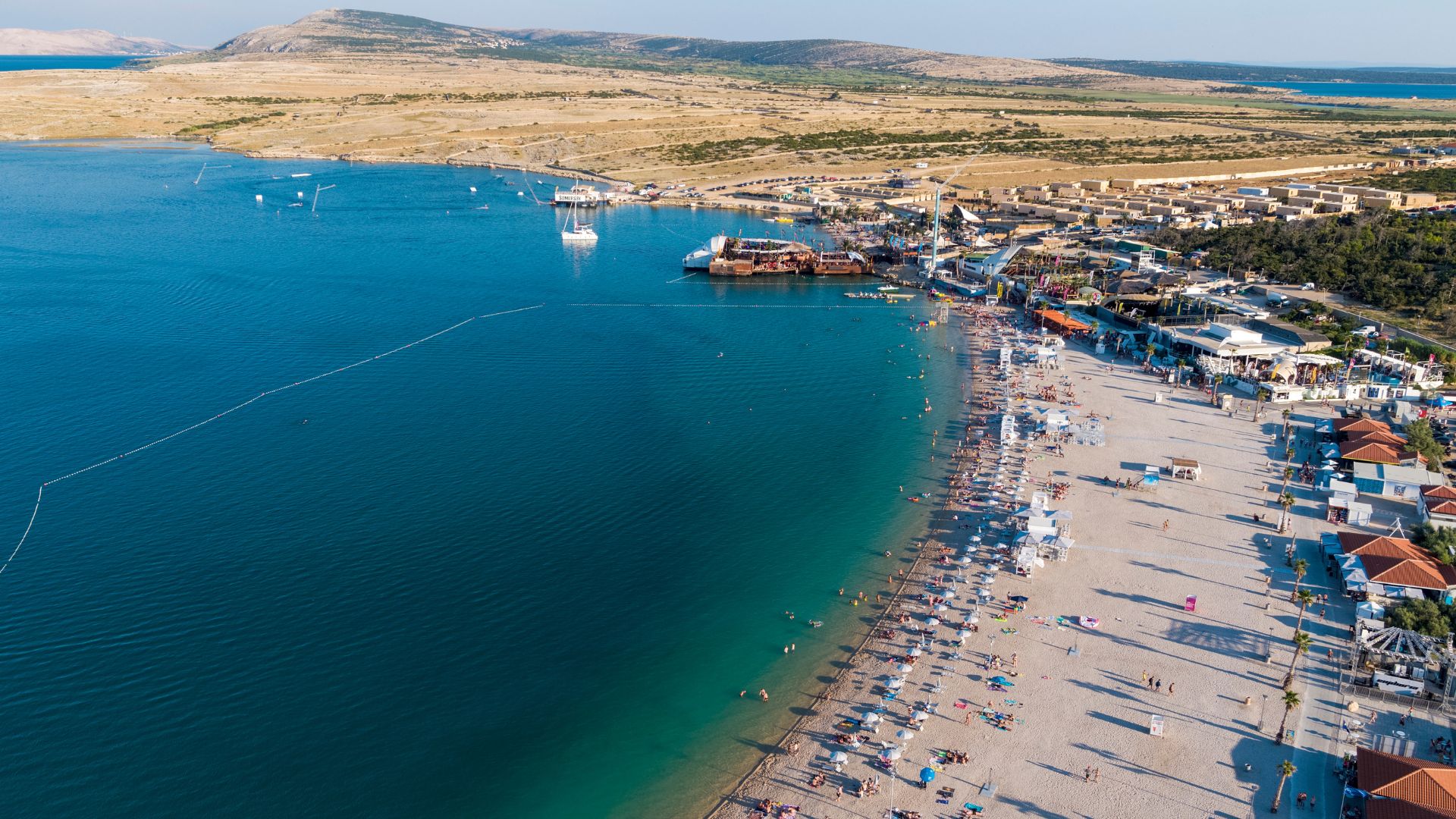 An aerial view of Zrće Beach in Croatia, showing a wide, sandy beach lined with buildings and umbrellas, a clear blue bay with boats, and arid hills in the background under a bright sky.