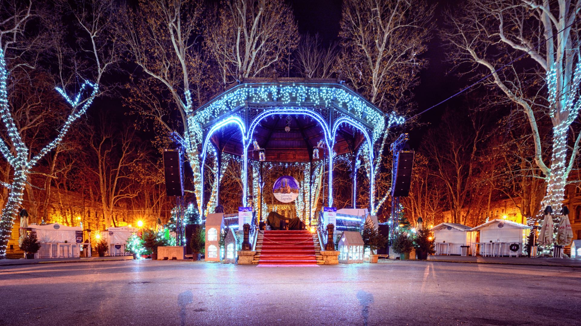 A beautifully decorated gazebo in a park at night, illuminated with blue and white Christmas lights, with trees also adorned with lights in the background, creating a festive winter scene.