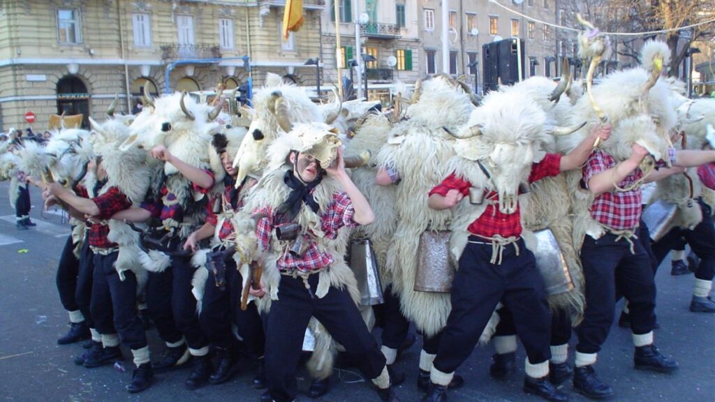 A group of Zvončari, traditional Croatian bellmen wearing sheepskin costumes, horned masks, and large bells, participate in a procession on a city street.