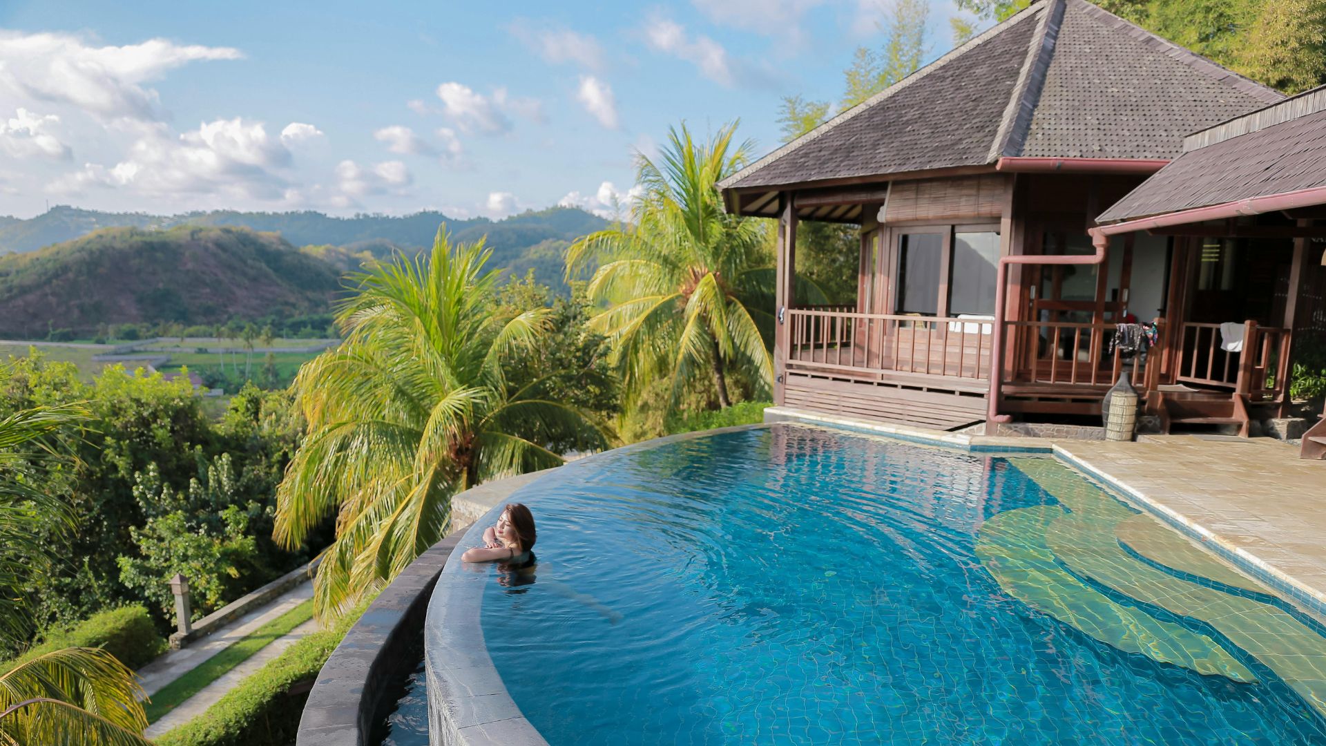A person in an infinity pool overlooking a lush green valley and hills in Bali, Indonesia.