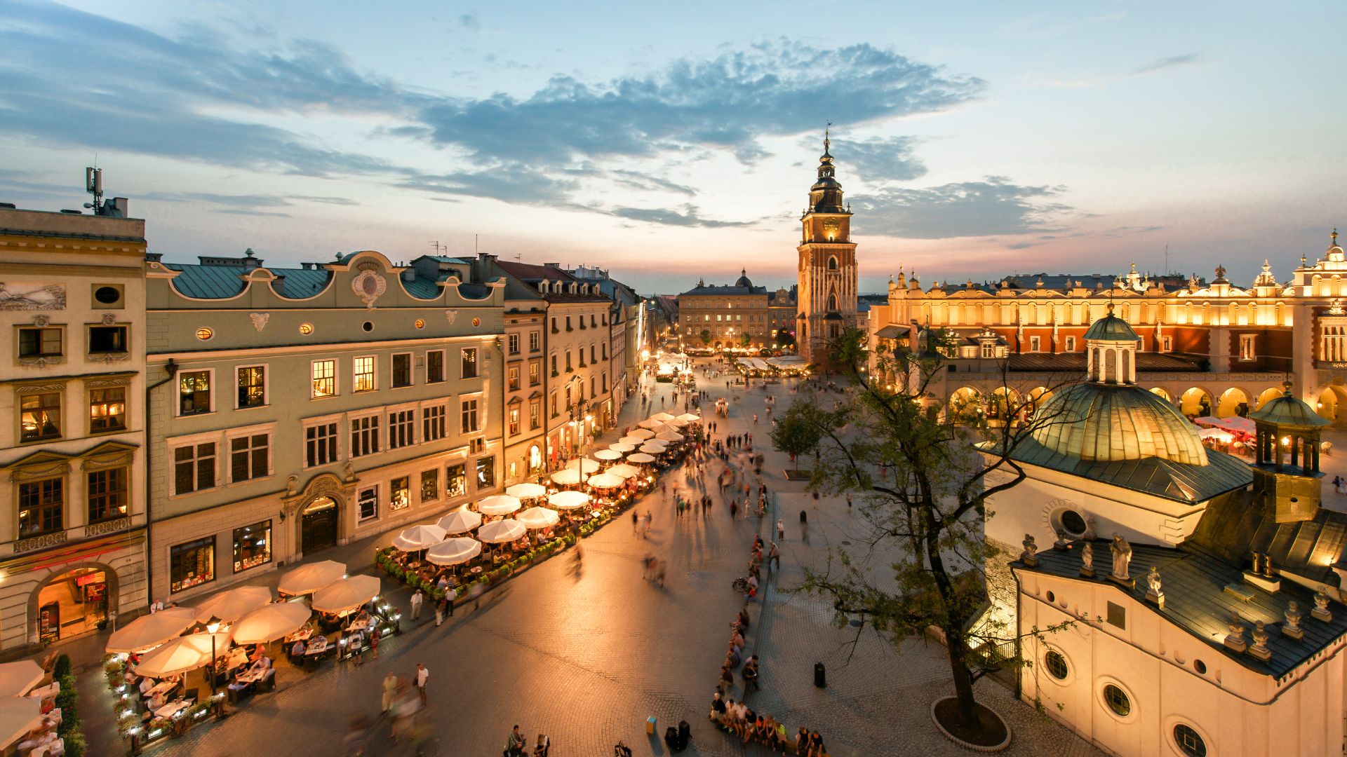 A vibrant, elevated view of the illuminated Main Market Square in Kraków, Poland, at dusk, showing historic buildings, outdoor cafes, and people walking.