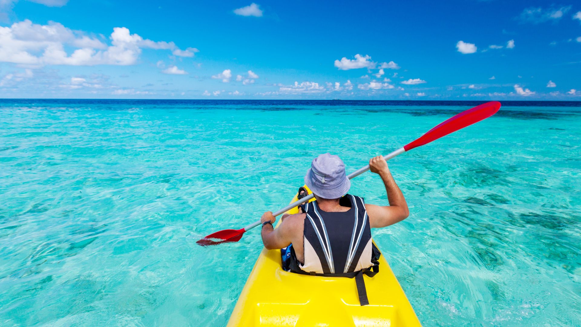 A person wearing a hat and life vest is kayaking in clear turquoise water under a blue sky with white clouds.