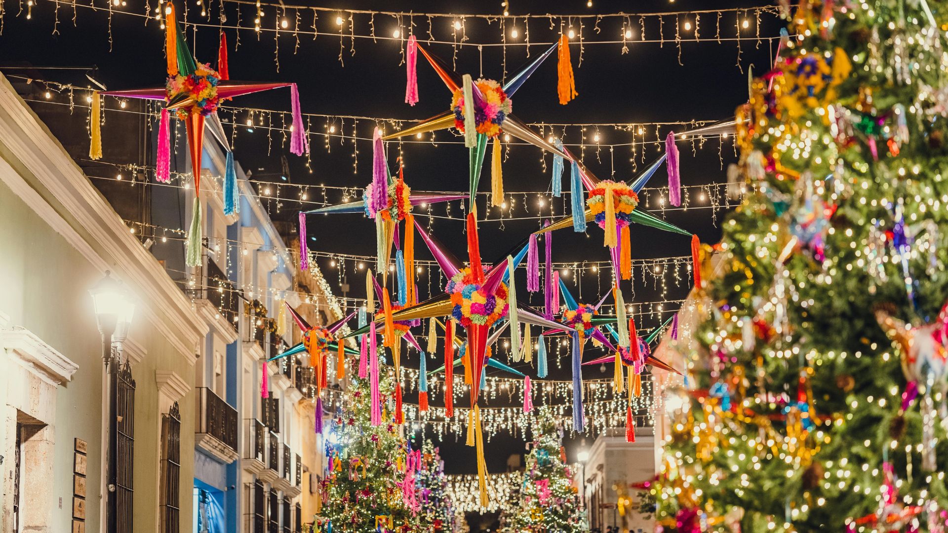 A street in Mexico decorated at night with colorful star-shaped piñatas and string lights hanging overhead between colonial buildings and a large Christmas tree.