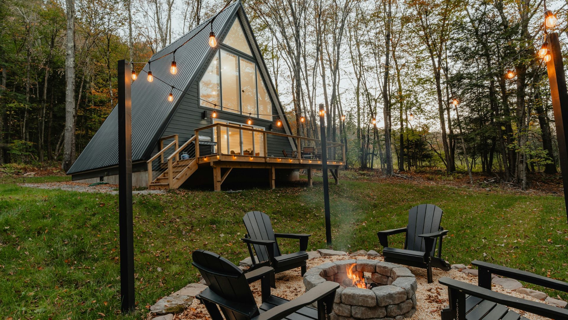 A modern black A-frame cabin with a large glass front and a lit deck sits in a wooded area at dusk, with a stone fire pit and black chairs in the foreground.