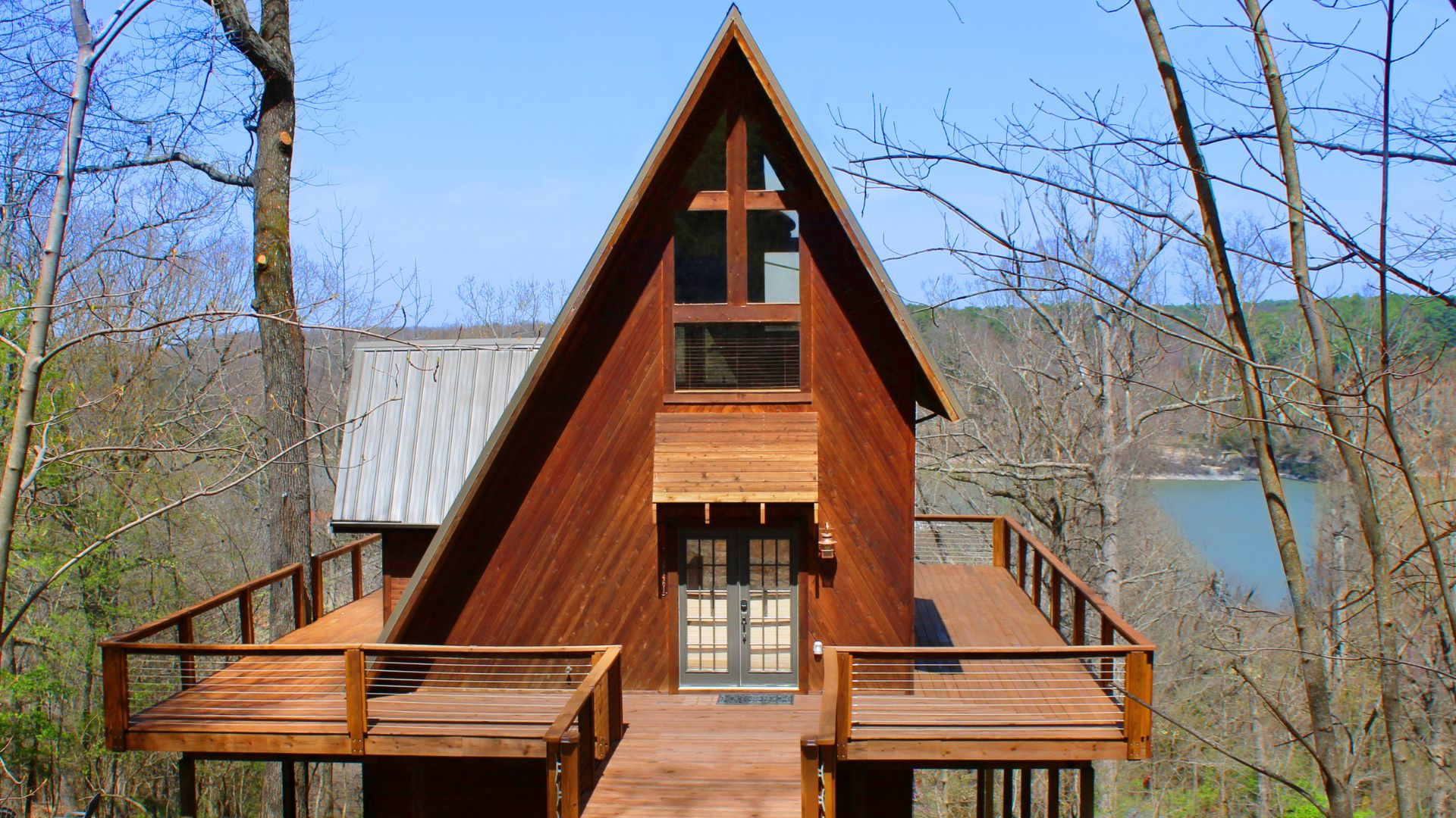 A wooden A-frame cabin with a large deck overlooking a calm lake or river, surrounded by trees with sparse foliage under a clear blue sky.