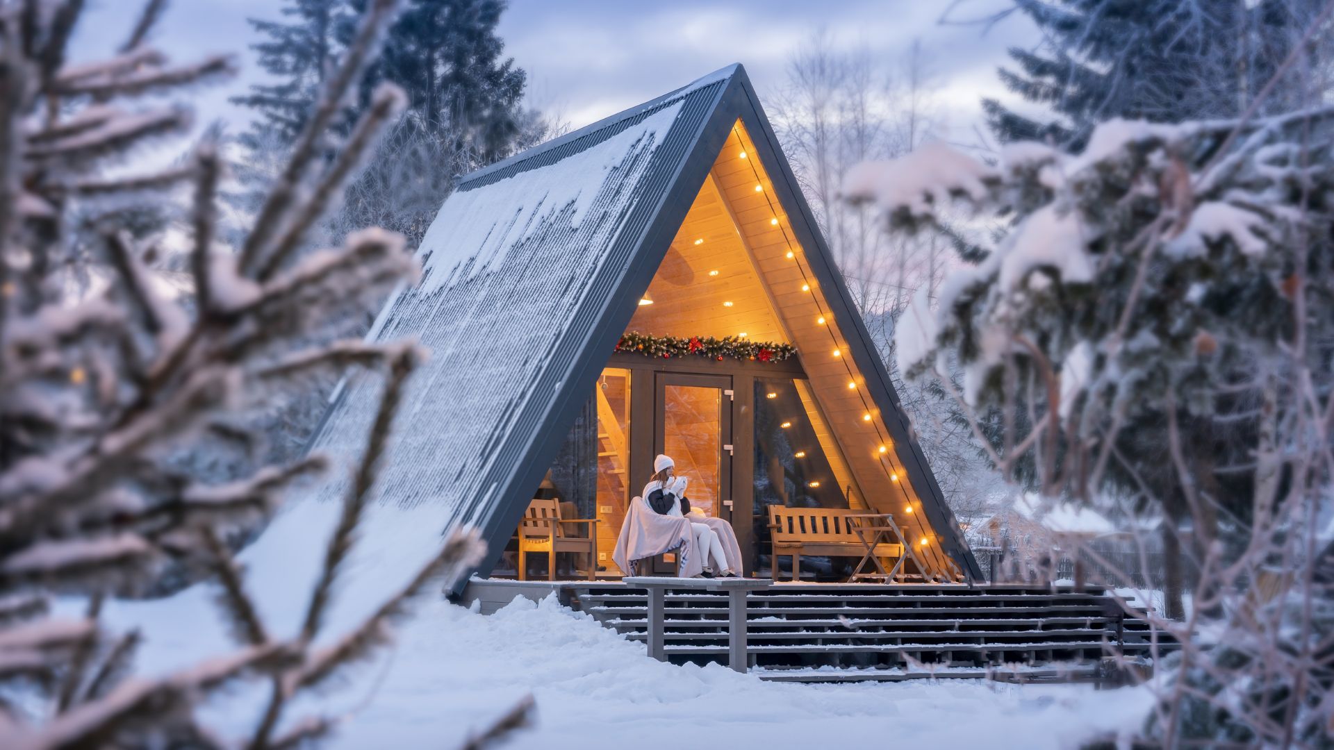 A cozy A-frame cabin nestled in a snow-covered winter forest, illuminated with warm lights and festive decorations on the porch.
