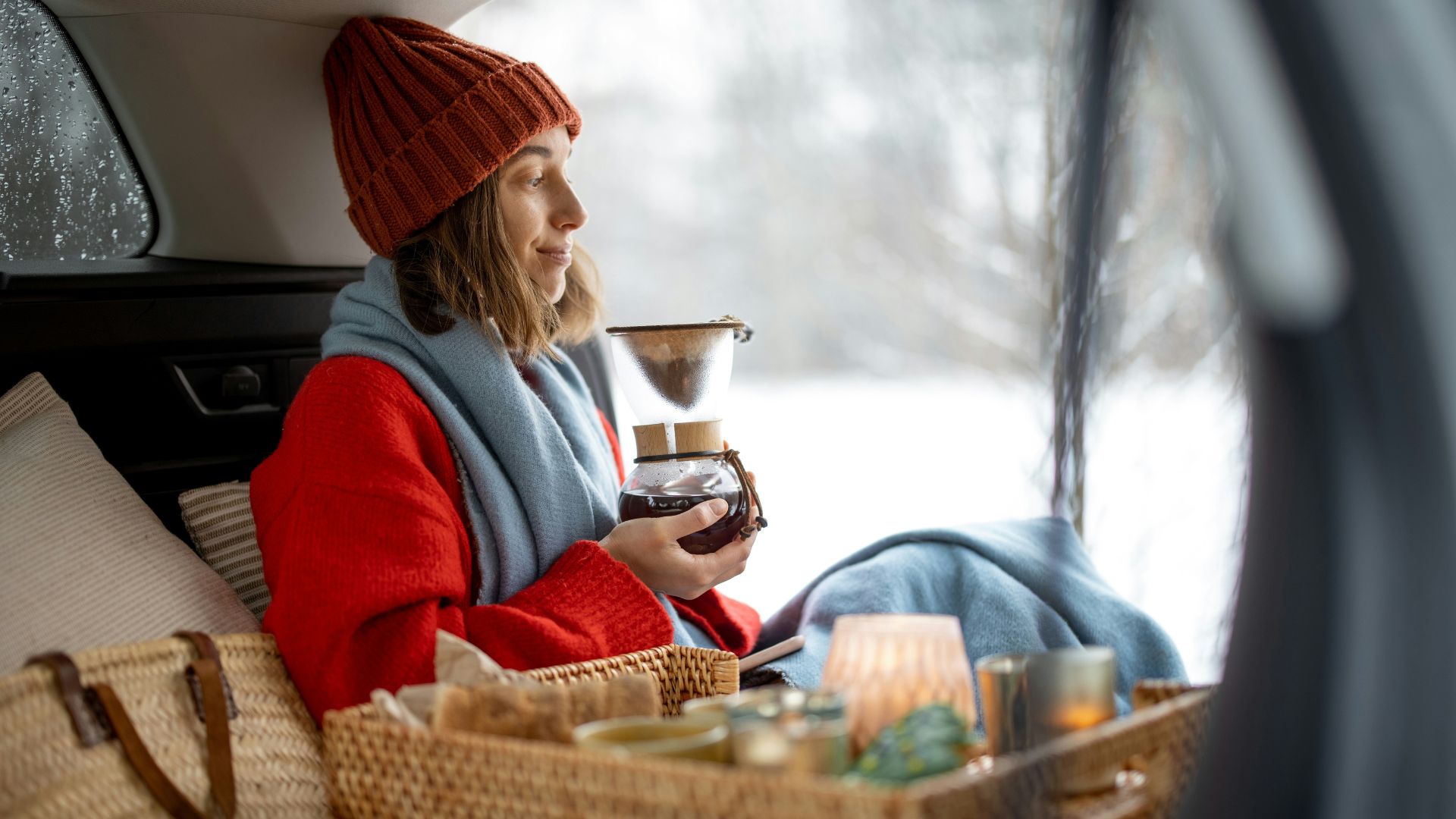 A womanwearing a red sweater and maroon bonnet holding a coffee maker inside her car on a snowy surroundings.